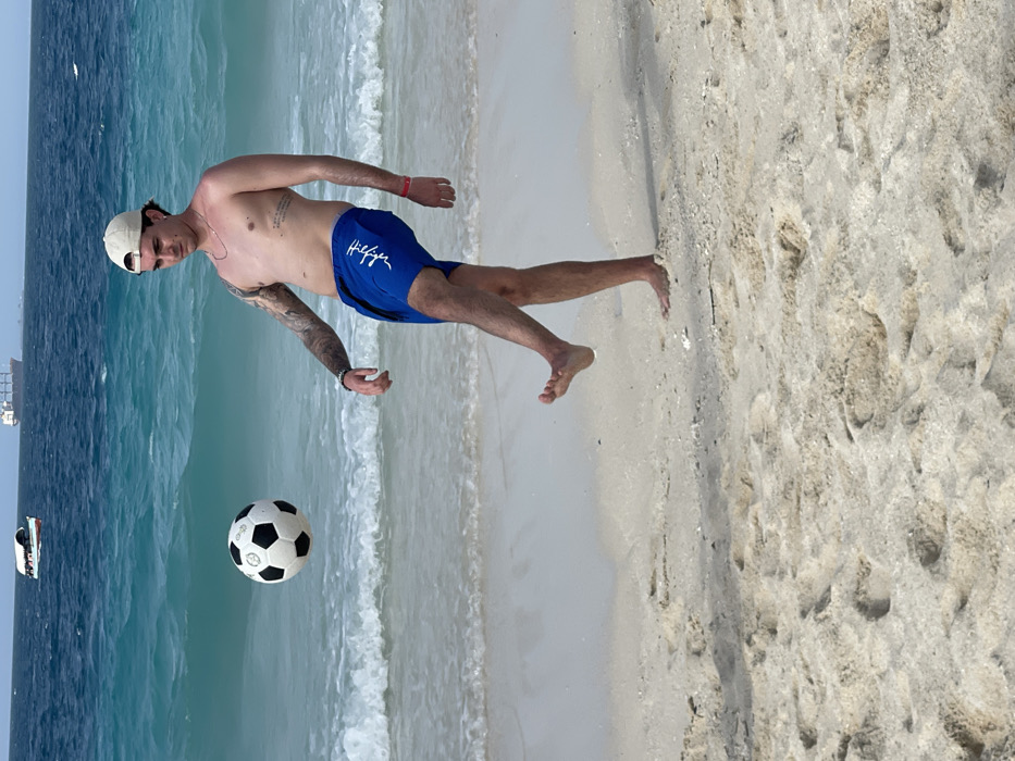 This image depicts a young adult male performing a beach soccer maneuver on a sandy shoreline, identified as being within the city of Zanzibar, Tanzania. The male, standing barefoot on the light-colored sand, is captured with his right leg raised, actively juggling a standard black and white soccer ball that is suspended mid-air just above his right foot. He is bare-chested, wearing blue swim shorts with white text, and a light-colored baseball cap. Visible tattoos adorn his left arm and the left side of his torso. He wears a thin chain necklace, a dark wristband on his left wrist, and a red wristband on his right. The immediate foreground shows sandy beach with distinct footprints. Behind the individual, the clear turquoise and deep blue ocean displays gentle waves breaking near the shore. In the distant background, a small white boat with a canopy and a larger cargo ship are visible on the horizon under a clear, bright blue sky.