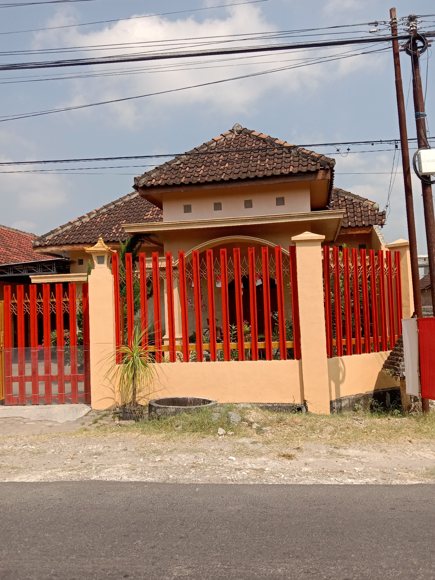 A single-story house with an orange-yellow facade and a terracotta tiled roof is visible on a sunny day. The house is partially obscured by a bright red metal fence with decorative scrollwork and vertical bars. A small, potted green plant with spiky leaves sits to the left of the fence. Behind the fence, lush green foliage is visible. The house is situated on a paved road, with a dirt and gravel area between the road and the fence. Utility poles and wires stretch across the pale blue sky, dotted with white clouds. The scene appears to be in Balongrejo, Indonesia.