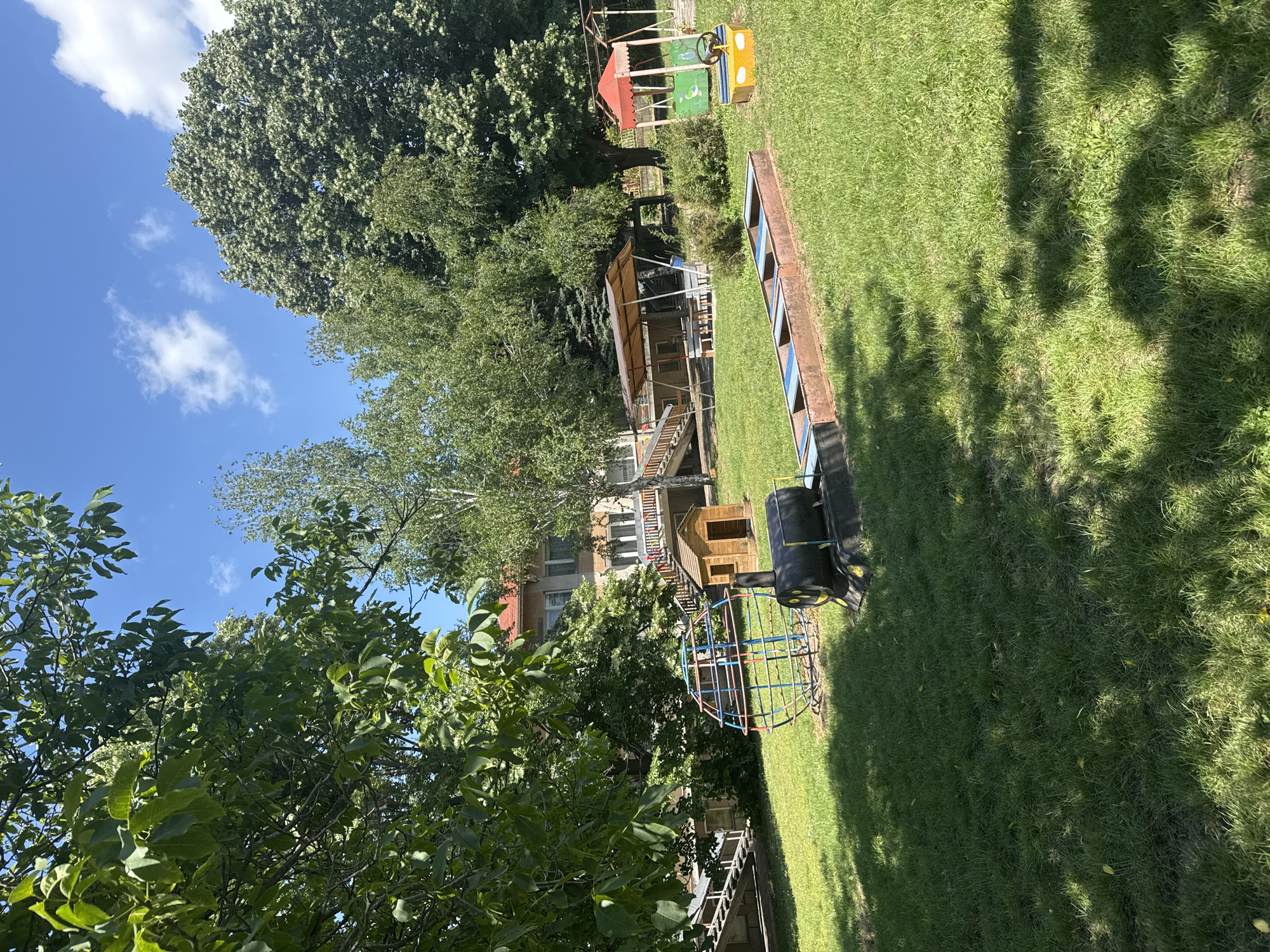 The image depicts an outdoor playground area under a clear blue sky with scattered white clouds, located in Basarbovo, Bulgaria. The scene is illuminated by strong sunlight, casting distinct shadows on the ground.
In the foreground, a vibrant green lawn occupies the lower half of the frame, showing variations in grass density and scattered leaf litter. Tree shadows are prominent across the grass in the lower half.
Central to the foreground is a spherical jungle gym constructed from red and blue metal bars. To its right, a black cylindrical object resembling a train engine, possibly made from repurposed barrels, features a yellow painted smiley face on its front. Extending to the right from this engine-like structure is a rectangular, multi-compartment play structure, possibly a sandbox or a series of shallow troughs, with blue inserts within a weathered wooden or metal frame, configured as train cars.
Further into the midground, to the right, is a small, bright yellow play structure with a red canopy and a steering wheel, resembling a miniature vehicle or swing frame. Directly behind it, a wooden gazebo or shelter with a sloped brown roof provides shade for a visible picnic table and benches. To the left of the gazebo, a small wooden playhouse with a brown gabled roof and a single window is situated on a slight elevation.
In the background, a multi-story building with a reddish-orange tiled roof is partially visible through and above dense green tree foliage. The building's facade appears to be light-colored, possibly painted stucco, and features multiple rectangular windows and visible balconies or railings. A set of wooden stairs leads up towards the building on the left side, with an additional concrete or stone staircase further left. The surrounding area is densely wooded with various broadleaf and coniferous trees, providing a lush green backdrop. No individuals are present in the scene.