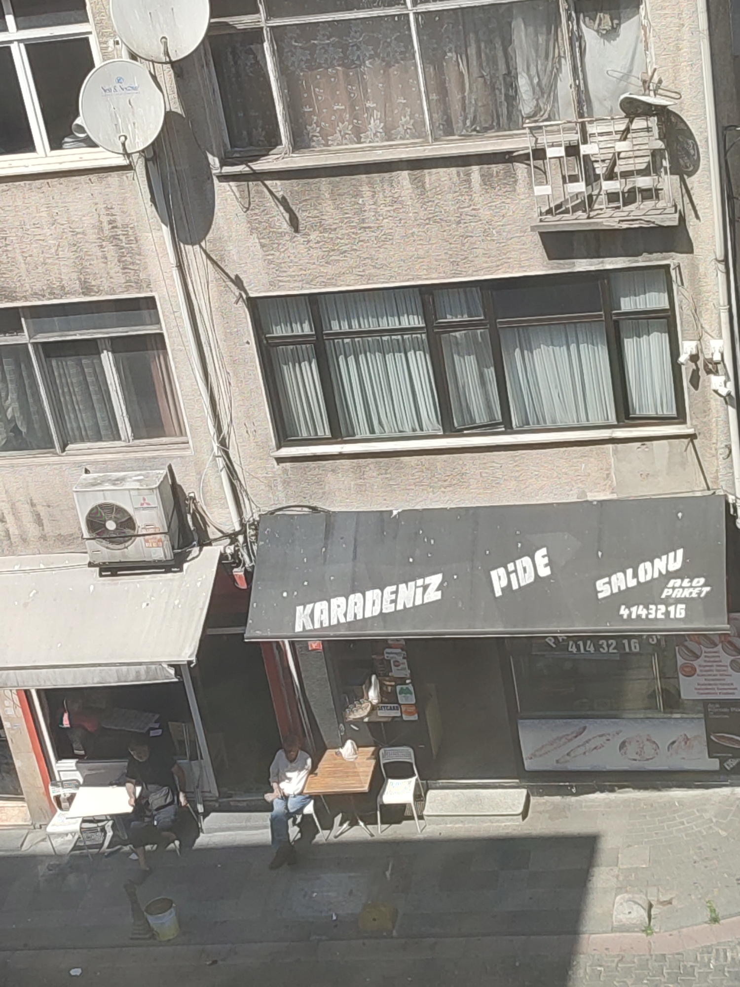 In the city of Kadıköy, Turkey, an outdoor scene unfolds with a view of a building's facade. The building features multiple stories with windows covered by white curtains. Several satellite dishes are attached to the upper floors. On the ground level, a business with a black awning displays white text that reads "KARADENİZ PİDE SALONU" and a phone number "4143216". Below the awning, a man in a white shirt and blue jeans sits at a table on the sidewalk, while another person, partially obscured, stands near an open doorway. A white air conditioning unit is mounted on the building wall above the awning. The ground is paved with cobblestones, and a yellow bucket is visible on the sidewalk.
