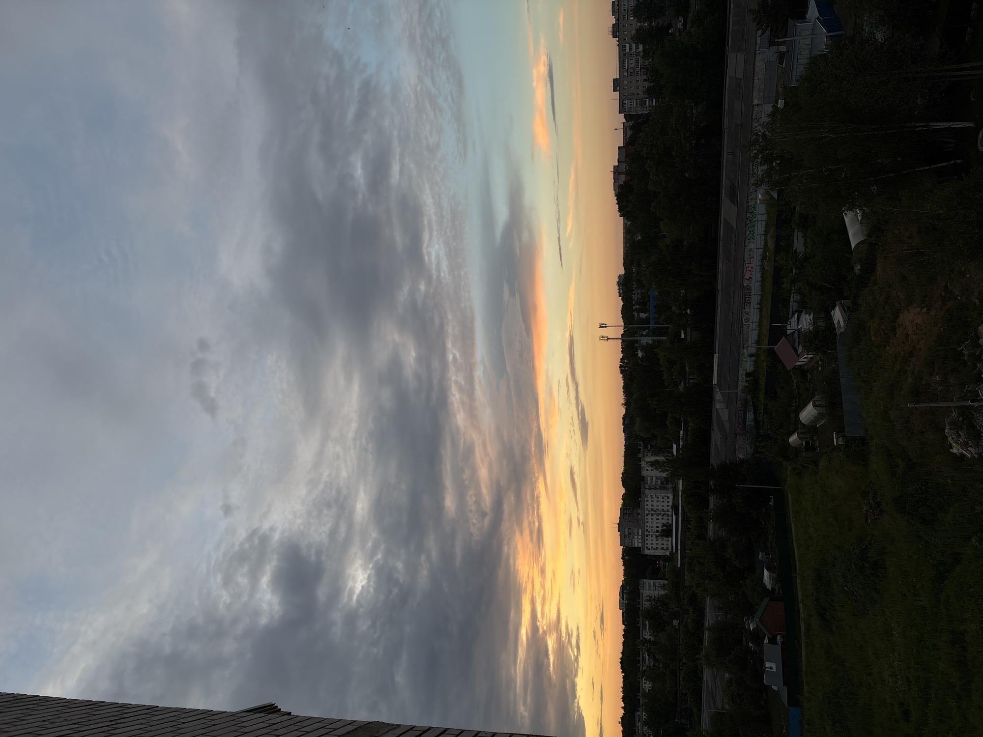 The image depicts a view of Chaikovsky, Russia, during what appears to be sunset or dawn. The sky is filled with dramatic clouds, transitioning from dark grays and blues at the top to vibrant oranges and yellows closer to the horizon. A brick building is partially visible on the far left of the frame, suggesting an urban or suburban setting. Below the sky, a densely forested area separates the sky from a collection of buildings, including multi-story apartment complexes and smaller structures. Some of these buildings appear to be industrial or commercial, with a long, low structure featuring graffiti. In the foreground, there is overgrown grass, some small sheds or greenhouses, and a dirt path, indicative of a more rural or undeveloped area on the outskirts of the city. No people are visible in the image.