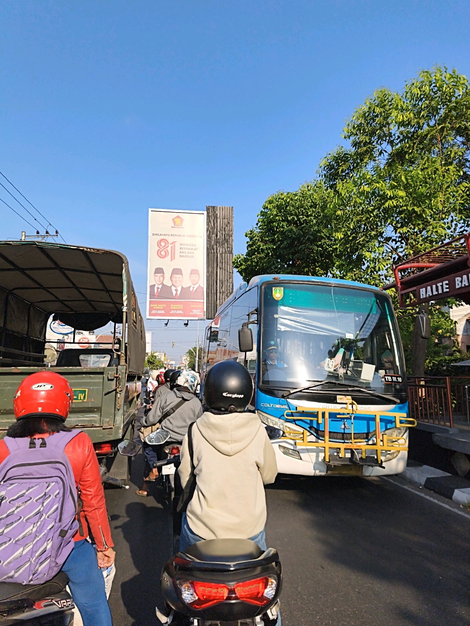 The image depicts a street scene in Surakarta, Indonesia, under a clear blue sky. A blue bus with yellow accents is prominently featured in the foreground, stopped in traffic. The bus has "COLT DIESEL" branding and a license plate ending in "10". To the left of the bus is a green military-style truck. Several motorcycles are visible, with riders wearing helmets. One rider in the immediate foreground wears a red helmet and a purple backpack. Another rider in the mid-ground wears a beige hooded sweatshirt and a black helmet. A large billboard advertising "DIRGAHAYU REPUBLIK INDONESIA 81" and featuring three men in suits is visible in the background, along with lush green trees. The street appears to be paved, and there is a sidewalk and a railing visible to the right. A sign that reads "HALTE BA" is also present. The overall impression is a busy urban environment during daylight hours.