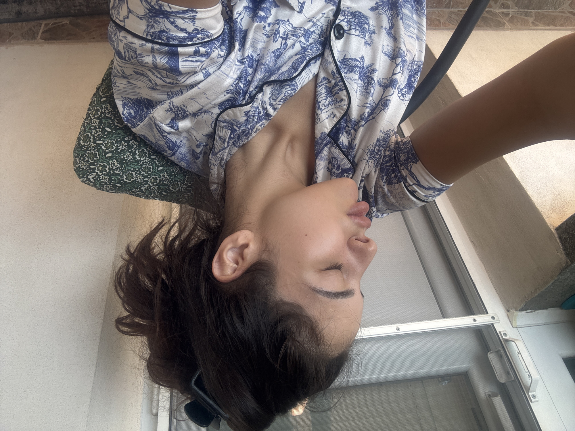 A young woman with dark hair pulled up in a messy bun and wearing blue and white patterned pajamas is seated on a chair, her head tilted back and eyes closed. Her lips are slightly parted, giving the impression she is about to speak or has just finished. Her left arm is visible, resting in her lap. The background shows a white wall and a window with a screen. The scene appears to be indoors, possibly a home or hotel room. There is no information to suggest the location is in Plovdiv, Bulgaria.