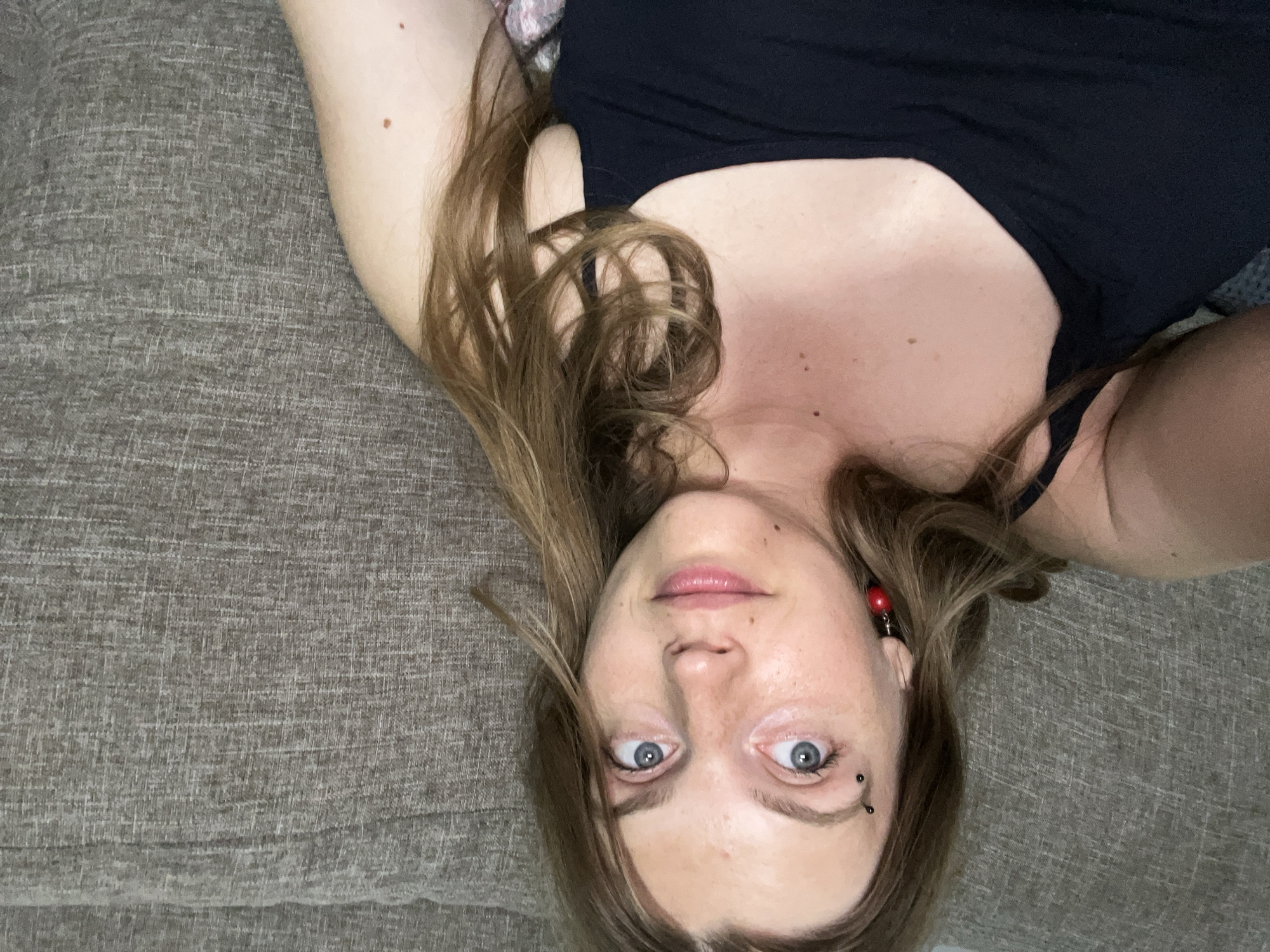 The image shows a close-up, high-angle selfie of a young Caucasian woman with light brown hair and blue eyes, wearing a black tank top and a red cherry-shaped earring. She has a piercing above her left eyebrow. She is lying on a textured gray couch. The lighting is natural and diffused. The scene is indoors, likely a living room. No specific location in Сеніцкі сельскі Савет, Belarus can be determined from the image.