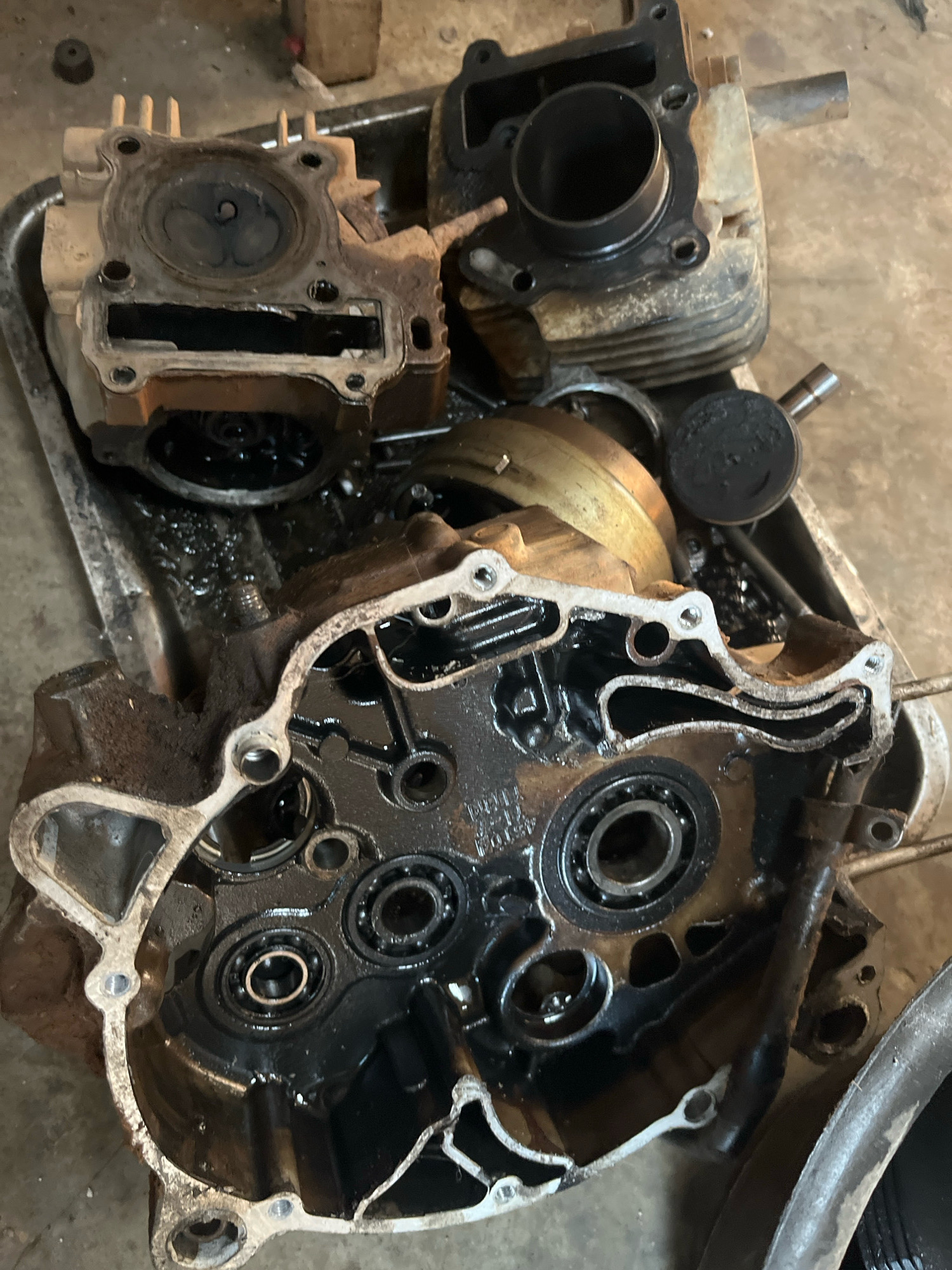 This is a close-up, high-angle photograph of disassembled motorcycle engine parts, likely in a workshop or garage setting in Kebumen, Indonesia. The components are dirty and greasy, suggesting they are either awaiting cleaning or are in the process of being repaired. The central focus is a large, open engine casing revealing multiple ball bearings submerged in dark oil. Above this, a cylinder head with visible valves and a piston, coated in soot, are laid out. Other engine components, including a cylinder and a part of a crankshaft, are also visible in the background. The overall environment appears utilitarian and suggests mechanical work is being undertaken.