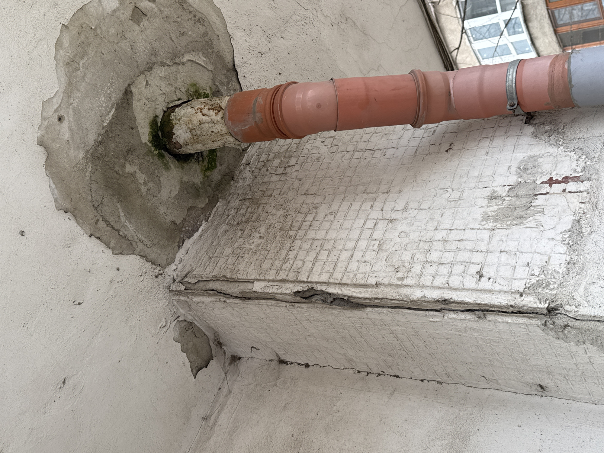 The image shows a section of an exterior wall, possibly of a building in Burgas, Bulgaria. A pipe, partially orange and gray, extends horizontally through a rough, circular opening in the wall. The opening's edges are chipped and fragmented. Green moss or algae growth is present around the pipe where it enters the wall. The wall's surface is cracked and appears to have a patterned texture, possibly from underlying tiles or blocks. There is evidence of peeling or flaking paint on the surface. The image is taken from a low angle, looking upwards at the wall and pipe. A window is visible in the upper right corner.
