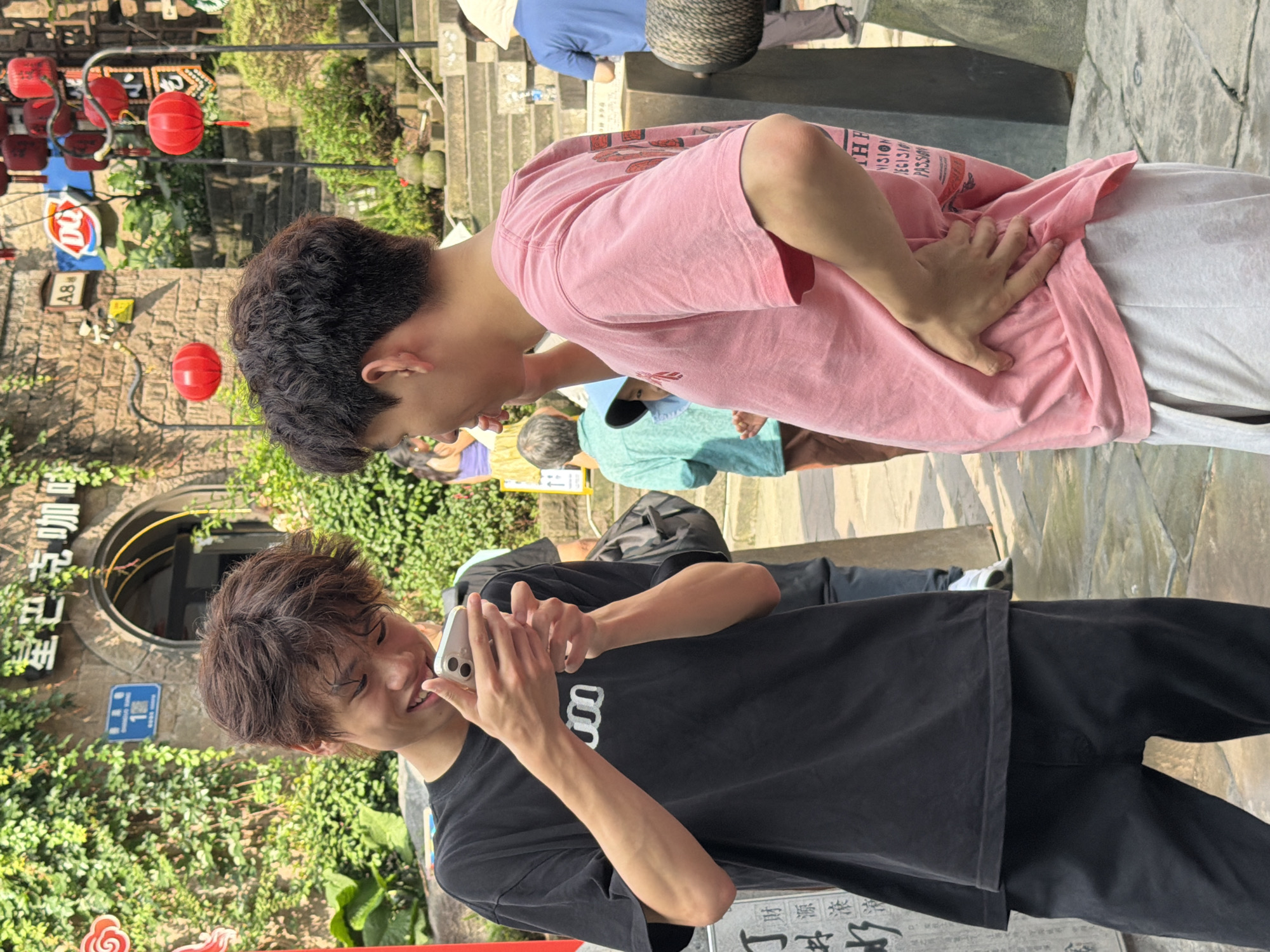 Two young men are standing on a stone-paved street in front of a building with greenery and lanterns. The man on the left, wearing a black t-shirt and black pants, is looking at a smartphone he holds in his hands and smiling. The man on the right, wearing a pink t-shirt and light gray shorts, is looking at the man on the left with his right hand on his hip. In the background, there are stairs, other people, and a building with Chinese characters and a Dairy Queen sign. The location is identified as Yuzhong District, China.