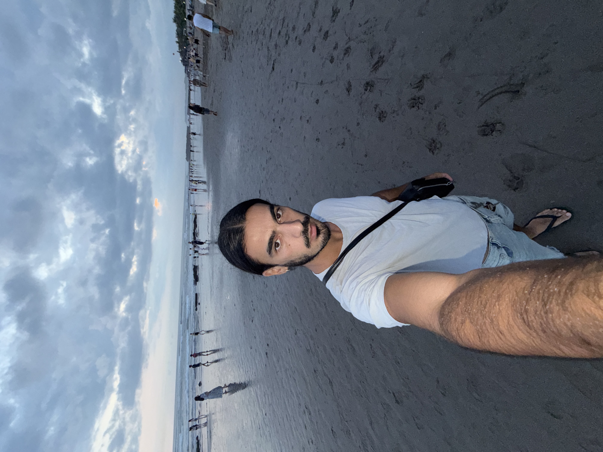 A man is taking a selfie on a beach in North Kuta, Indonesia. He is wearing a white t-shirt, light-colored shorts, and flip-flops. He has a black bag slung across his shoulder. The beach is sandy with dark sand and footprints. In the background, there is a crowd of people walking along the shoreline. The sky is overcast with clouds.