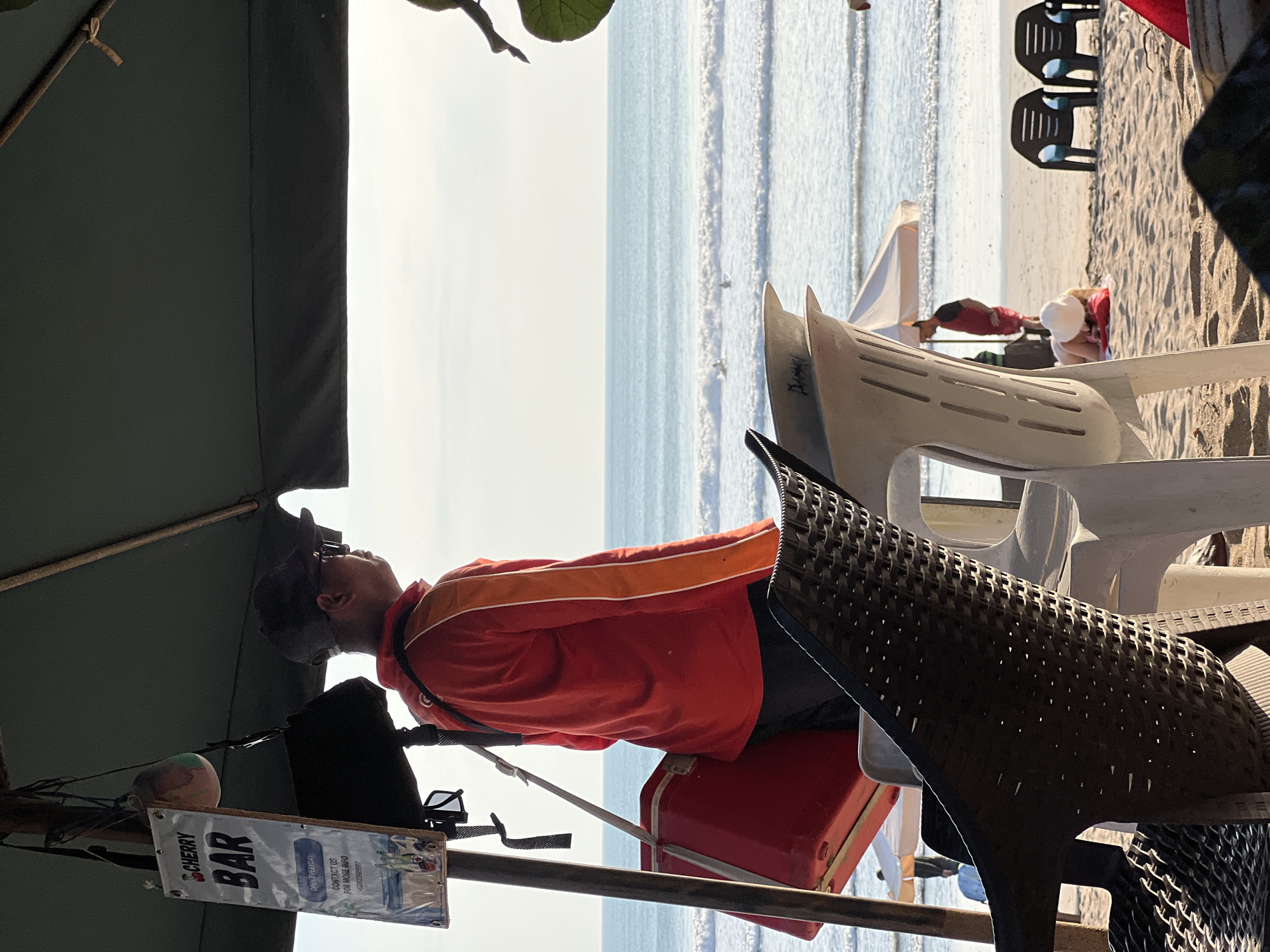 This is a medium shot of a beach scene in Kuta, Indonesia. In the foreground, the woven texture of dark and white plastic chairs is visible. Behind the chairs, a person wearing a red long-sleeved shirt with orange stripes on the sleeves and dark pants stands facing the ocean. They are wearing sunglasses and a baseball cap. A red cooler is slung over their shoulder. A sign for "CHERRY BAR" hangs on a pole near the person. The ocean with gentle waves is in the background, and a few surfers are visible in the water. The sky is bright and slightly overcast. The scene suggests a casual beach setting, possibly a bar or service area by the sea.