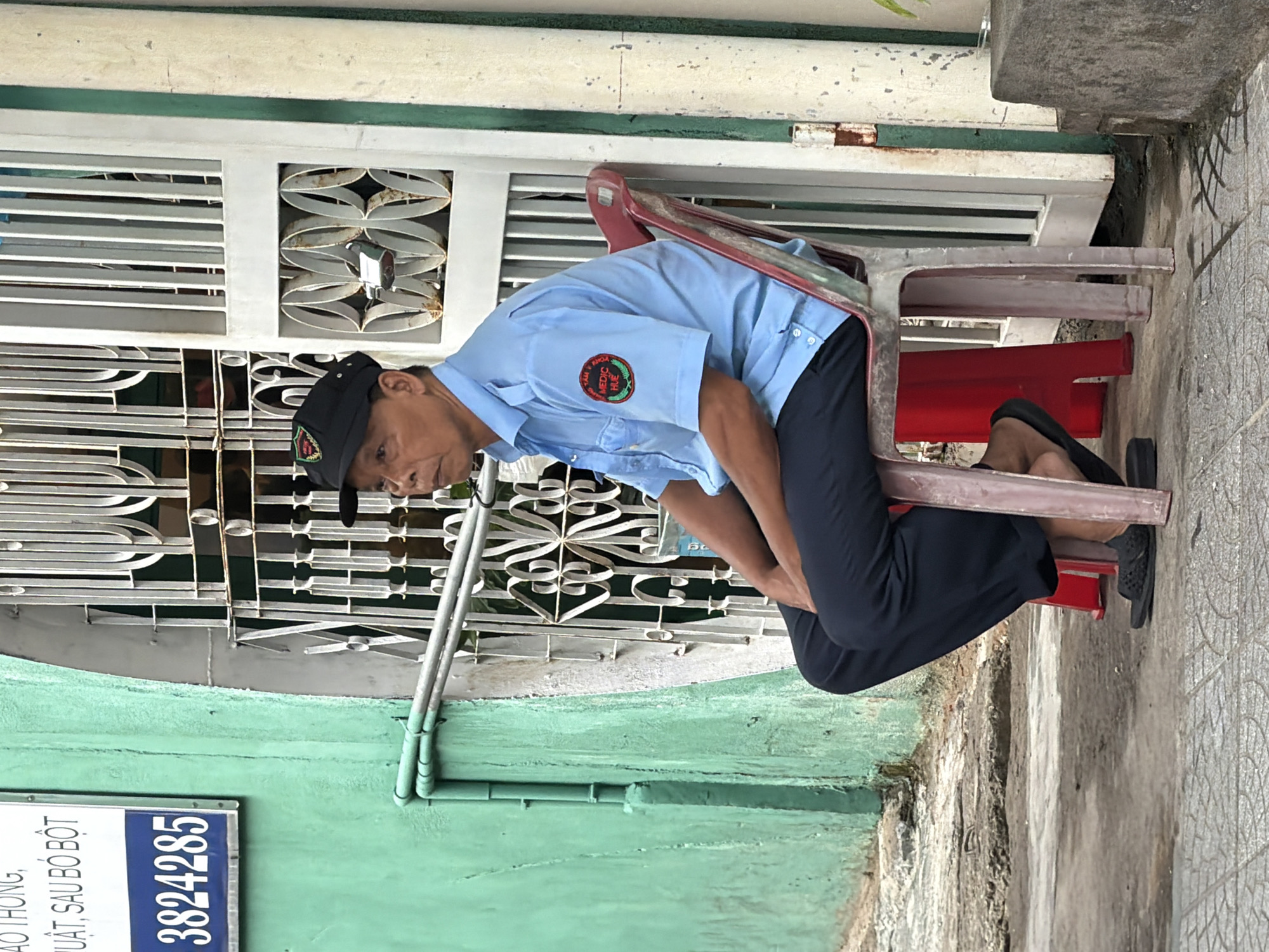 A middle-aged man wearing a light blue short-sleeved collared shirt with a red and green circular patch on the left sleeve, dark pants, and black sandals sits on a red plastic chair. He wears a black baseball cap with a similar circular patch. He is facing to his left, looking forward. The man is seated on a sidewalk in front of a building with a light green painted wall on the left side of the image and a white metal gate on the right side. A blue sign with white text and the number "3824285" is attached to the green wall. The gate has an ornate design and a padlock. The ground is paved with concrete and tiles.