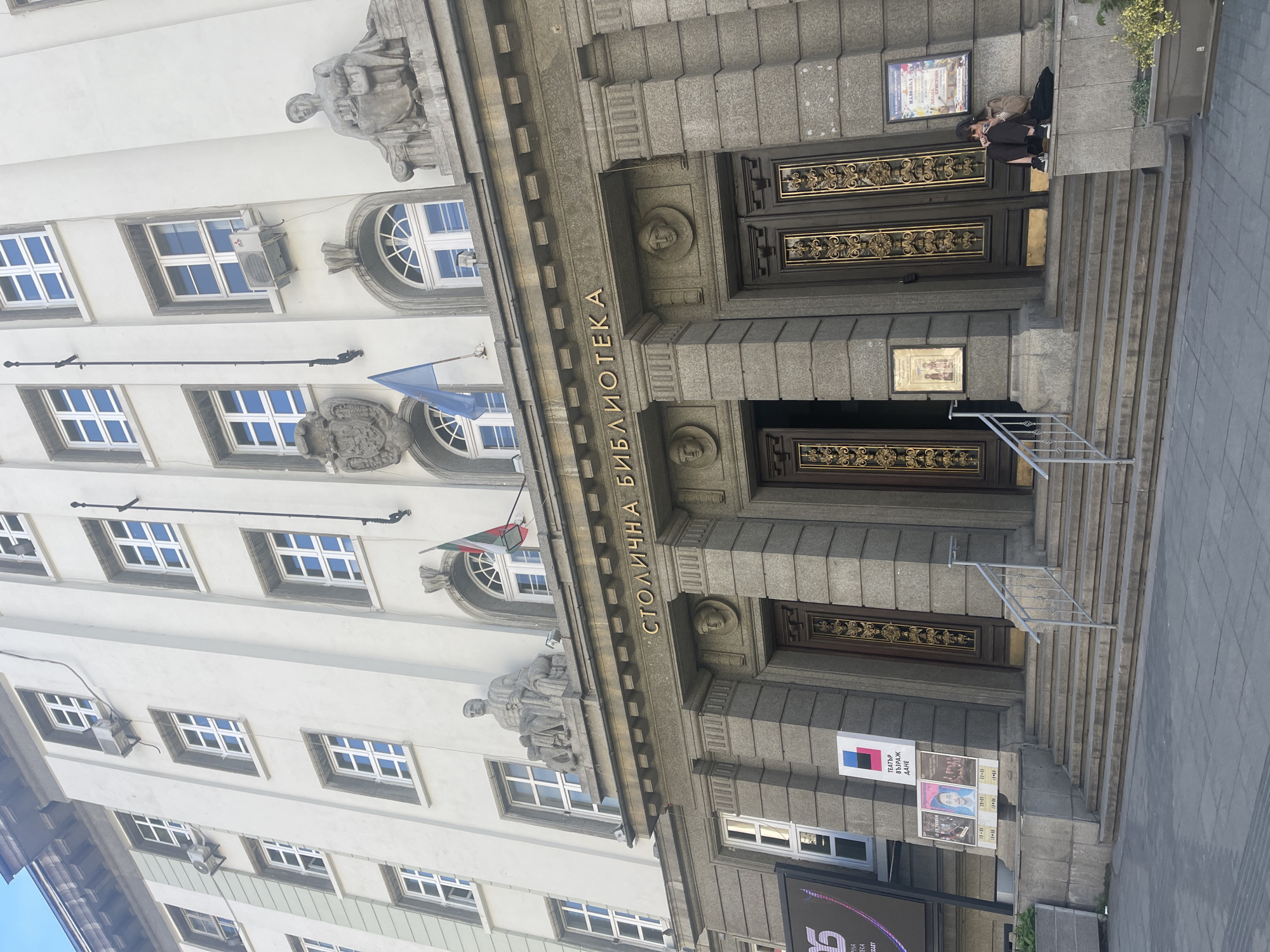 The image depicts the facade of the Sofia Municipal Library in Sofia, Bulgaria. The architectural style is classical with ornate detailing. The building is made of light-colored stone, possibly granite or a similar material. Above the entrance, the building's name, "Столична библиотека" ("Stolichna Biblioteka" - Sofia Municipal Library) is vertically inscribed. There are statues of figures in repose above the entrance. The doors are dark wood with decorative gold accents.
A person is sitting on the steps leading up to the entrance. There are posters and signs displayed near the entrance, including those for a theater ("Театър Възраждане"). Flags are displayed on the building's facade. The sky is visible between the windows, suggesting a clear day. Windows are on the facade of the building.