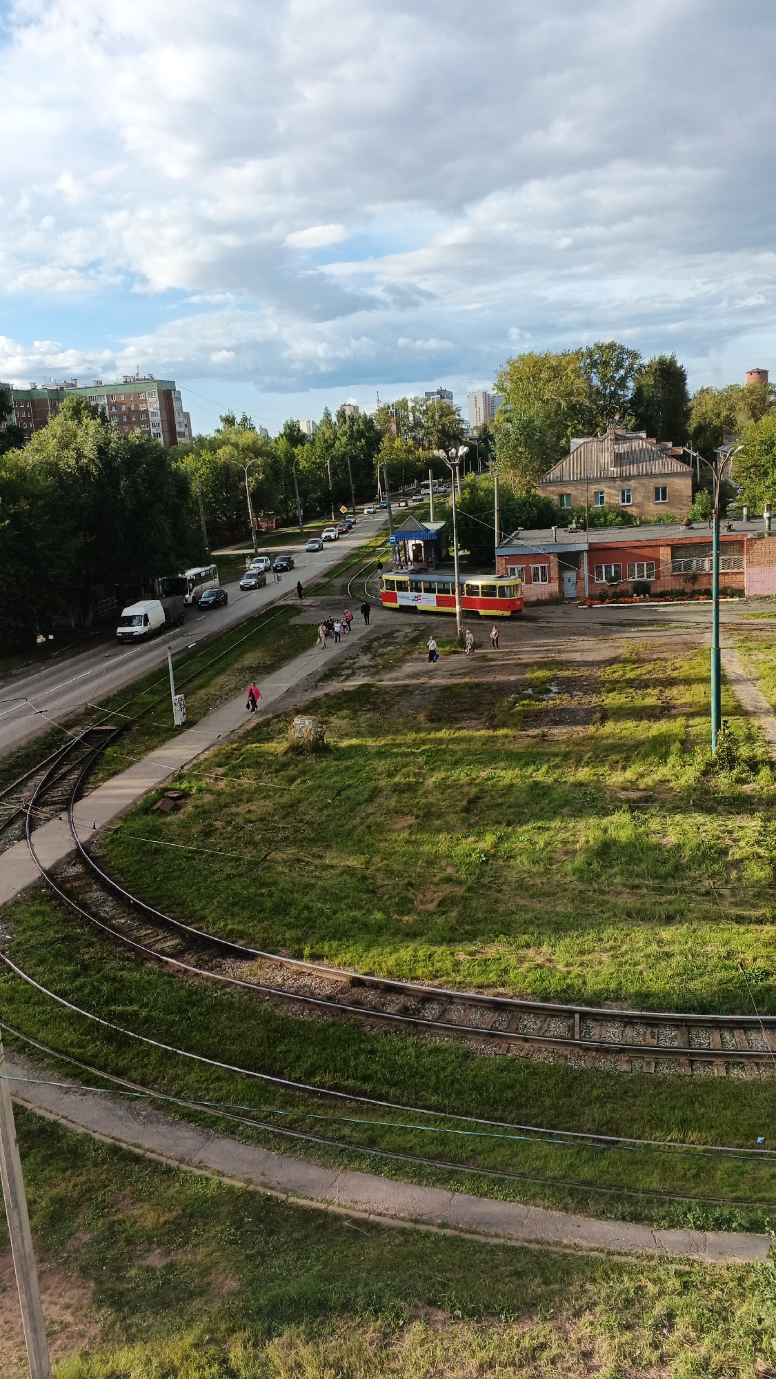 The image depicts a street scene in Izhevsk, Russia, with tram tracks prominently featured in the foreground. Several trams, one red and yellow and another red with yellow accents, are visible. The red and yellow tram is stopped at what appears to be a designated tram stop, where a group of pedestrians is gathered. Another red tram with yellow accents is also present, possibly at a depot or another stop.
In the background, a two-story brick building with multiple windows and a darker roof stands to the right of the tram stop. Adjacent to it is a smaller, single-story brick structure, possibly part of the same complex. A tall, green metal pole, likely a utility pole or street light, is situated in front of the brick buildings. Further in the background, residential buildings with multiple stories are visible, surrounded by lush green trees. A road runs alongside the tram tracks, with several cars and a white van present. The sky is partly cloudy, with patches of blue sky interspersed with white and gray clouds. The overall scene is one of an active urban environment with public transportation and residential areas.
