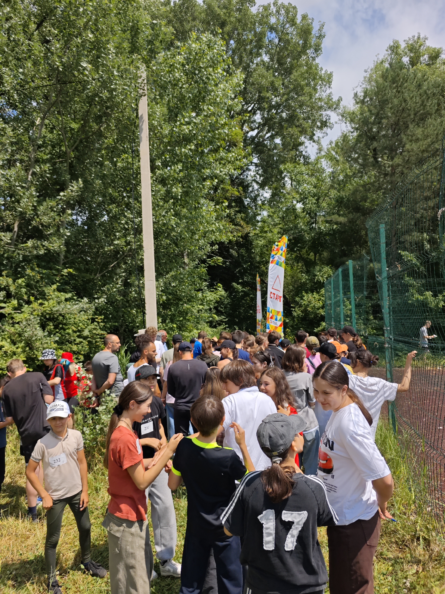 An outdoor gathering of people, likely for a sporting event, is depicted in an image. The scene is set amidst trees and vegetation, with a fence and a tall pole also visible. A group of individuals, appearing to be of various ages, are standing and interacting. Some are wearing athletic attire, and several have bib numbers pinned to their clothing, suggesting participation in a race or competition. Two prominent flags, one with the word "СТАРТ" (START in Russian) printed on it, indicate the event's commencement point. The overall atmosphere seems casual and communal, with people engaged in conversation and preparation. The location within Cherkessk, Russia, is not directly identifiable from the image's visual cues alone.