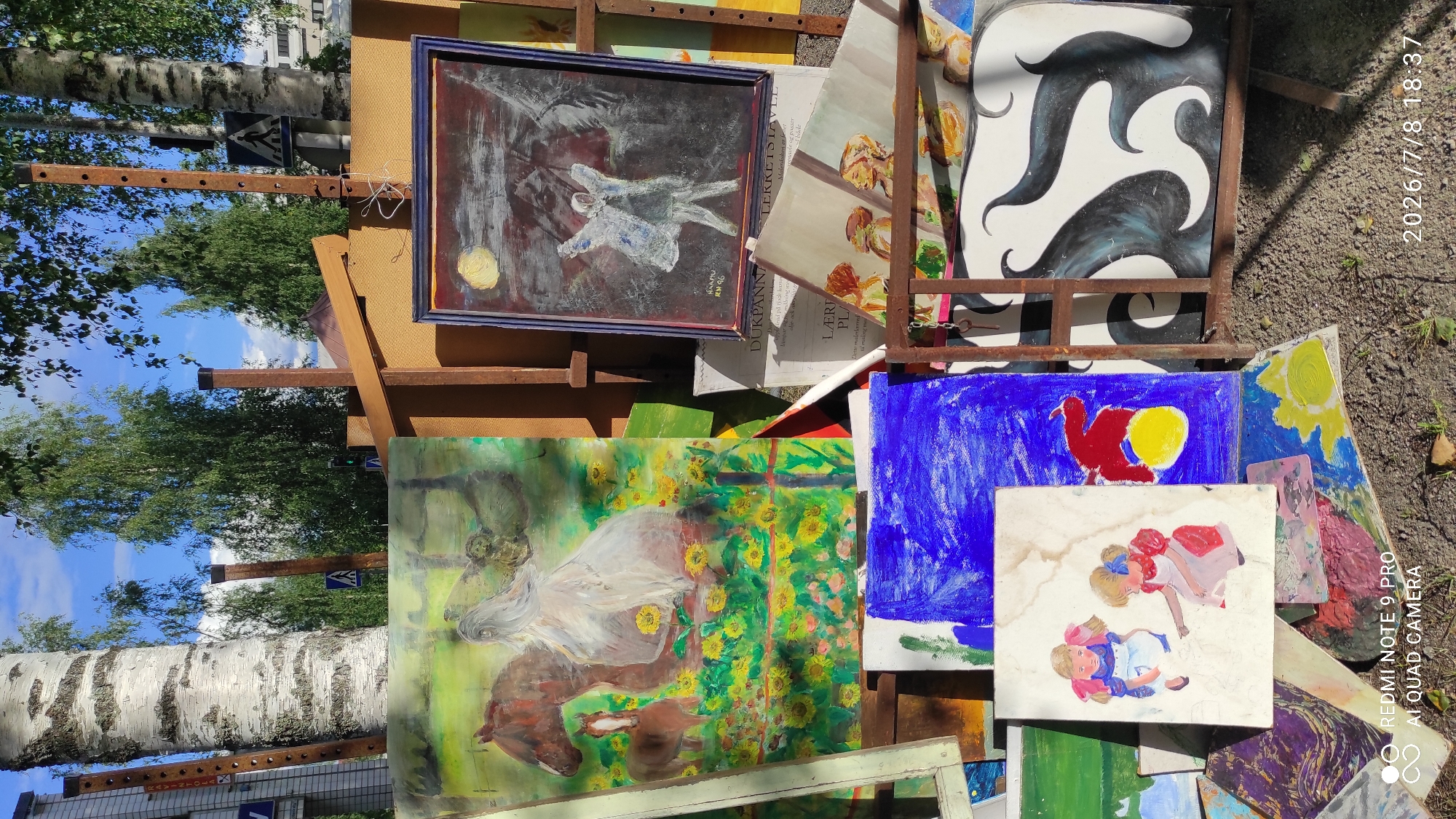 This media file captures an outdoor art display, likely a market or exhibition, in Iisalmi, Finland. The scene is a vibrant, slightly chaotic arrangement of paintings propped up on stands and stacked on the ground. The artworks vary in style and subject matter, including abstract pieces with bold black and white swirls, a painting of a horse amidst sunflowers, a depiction of two figures, and a naive art piece featuring a red bird against a blue background. There are also framed paintings, one of which shows a ghostly figure with a yellow moon.
The setting is outdoors, with lush green trees and a bright blue sky visible in the background, suggesting a sunny day. The time indicated on the image is July 8, 2026, at 18:37, implying it's late afternoon or early evening. The ground is covered in dirt and leaves, reinforcing the outdoor nature of the display. There are also some wooden structures and metal framing holding the artwork.
Visible text elements include partial words and phrases that appear to be from labels or signage associated with the art, such as "NAAMU," "RM 96," "DUKPANN," "LÆRU PL," "Malerduken er festet," and "LEKKETSIAVLE." A watermark at the bottom right corner reads "REDMI NOTE 9 PRO AI QUAD CAMERA." The overall impression is one of a lively and creative artistic gathering set against a natural backdrop.