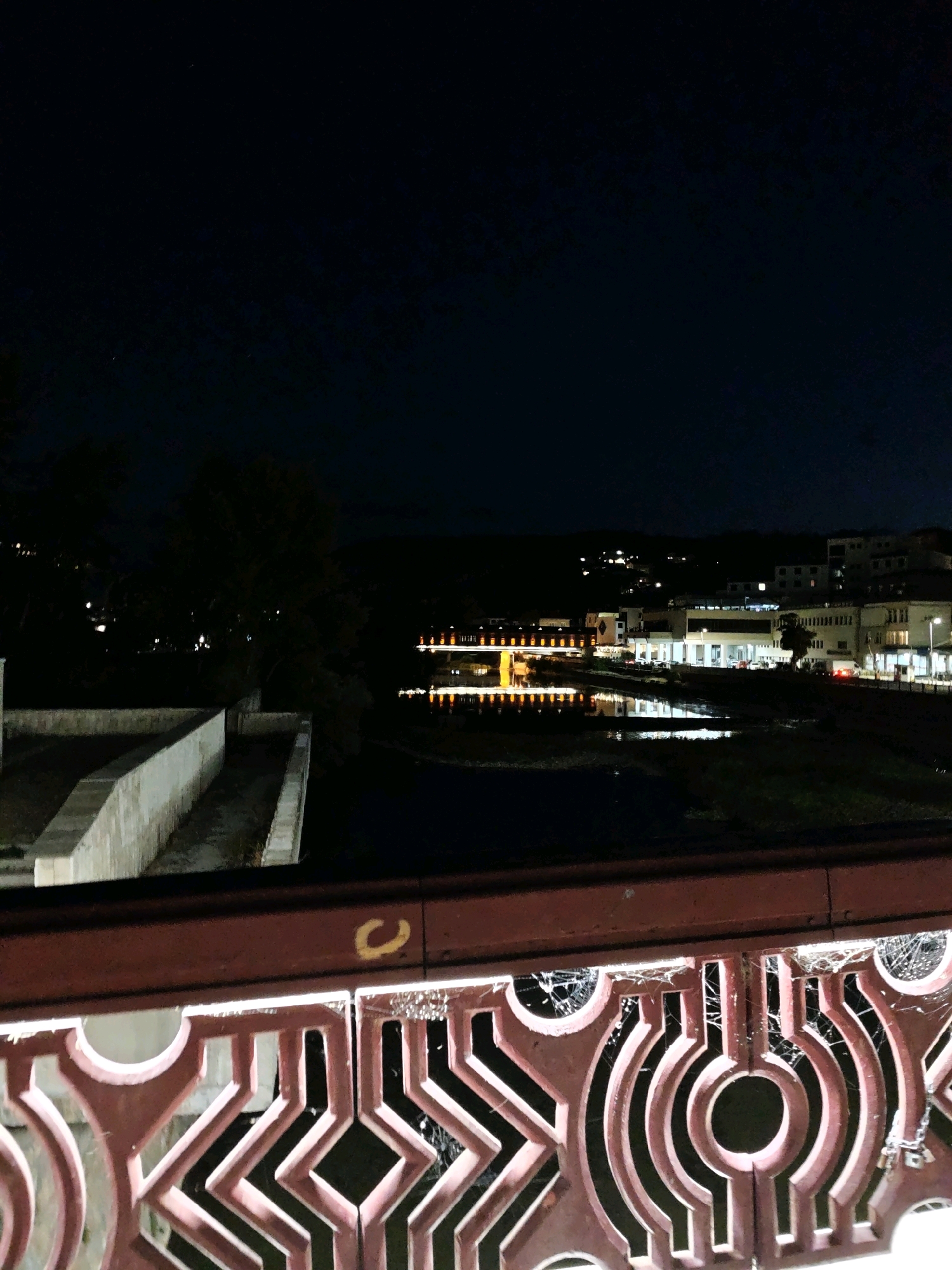 This is a night-time photograph taken from a bridge in Lovech, Bulgaria, overlooking the Osam River. In the foreground, the ornate red railing of the bridge is visible, adorned with intricate geometric patterns and some visible spiderwebs. The railing has a somewhat weathered appearance.
Beyond the bridge, the dark water of the river reflects the lights of buildings on the opposite bank. These buildings appear to be a mix of modern structures with illuminated windows, suggesting a commercial or residential area. Some of these buildings are situated on a hillside, with scattered lights indicating their presence. A distinct yellow marking, possibly a spray-painted "C", is visible on the upper horizontal beam of the bridge railing. The sky above is dark, with a few faint stars discernible. The overall impression is a quiet, urban night scene along the river.