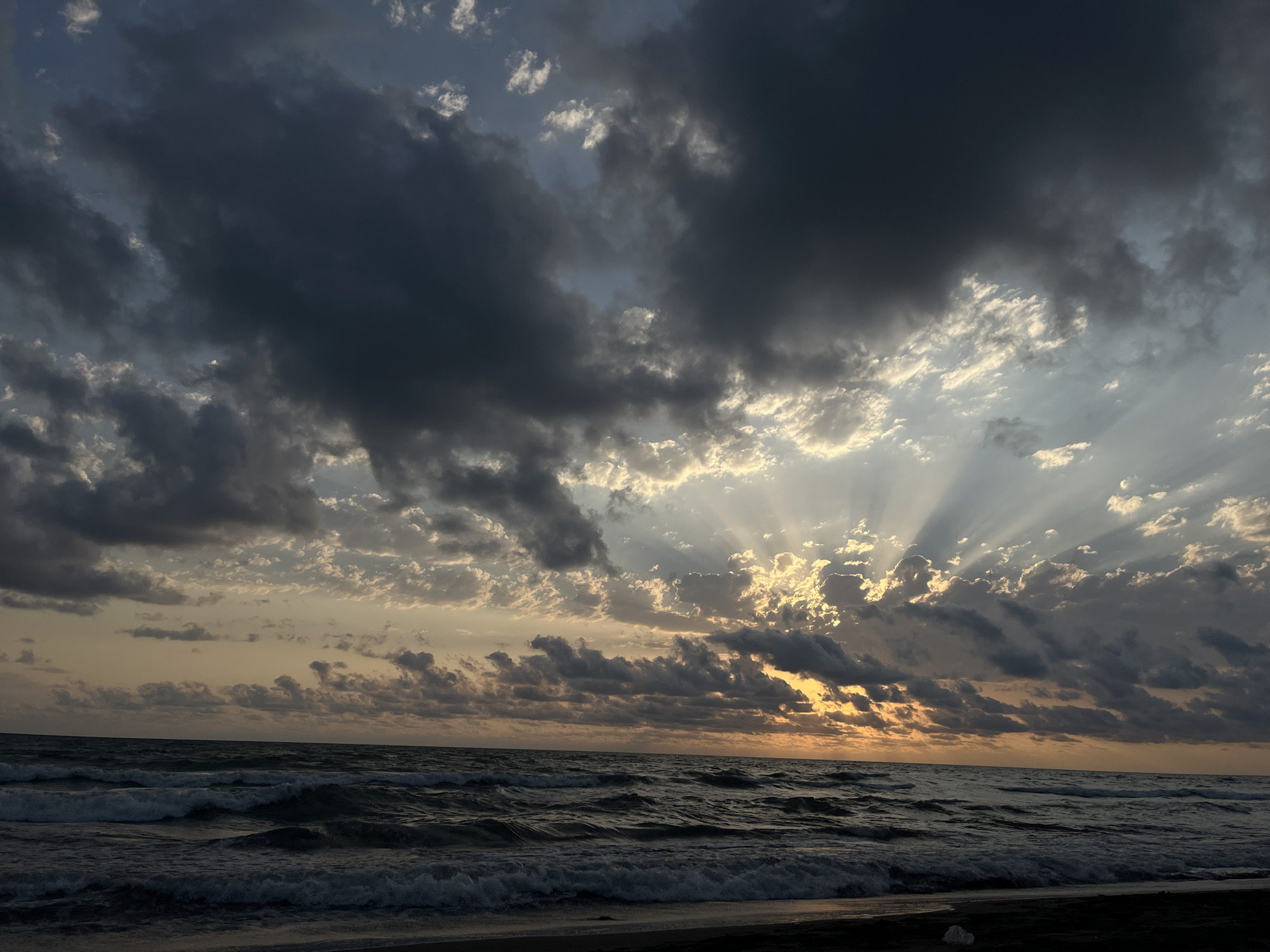 The image depicts a dramatic sunset over the Black Sea near the village of Supsa in Georgia. The sky is filled with dark, ominous clouds, but shafts of sunlight break through, creating a striking contrast. The sun itself is low on the horizon, casting a warm, orange glow. The sea is rough, with waves crashing onto the dark sand beach in the foreground. The overall mood is one of power and beauty.