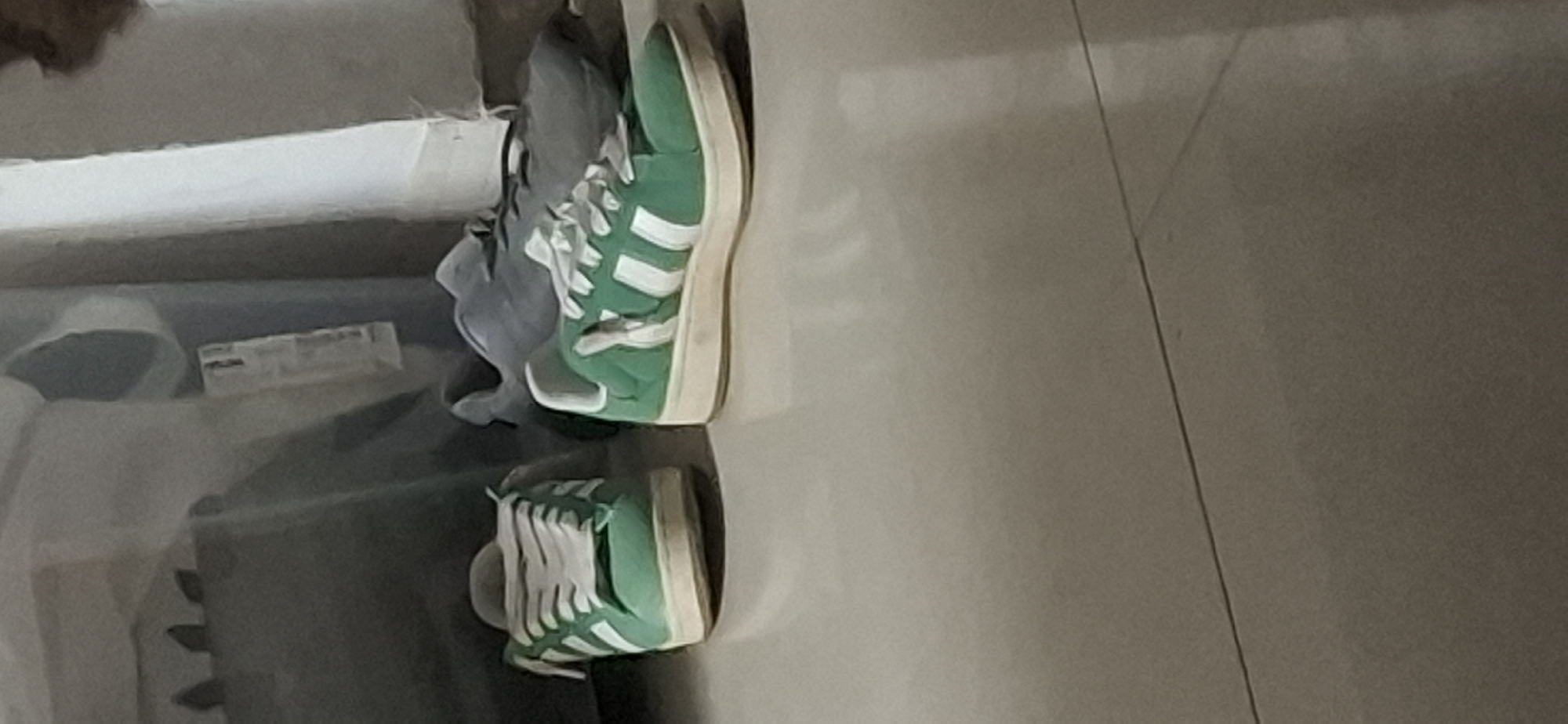 Two green Adidas sneakers with white stripes and laces are on a tiled floor. One sneaker is to the left and appears smaller, while the other is to the right and appears larger. Behind the sneakers, a large gray storage container or bin is visible, along with a lighter colored storage bin or shelving unit. The environment suggests an indoor space, possibly a home or storage area, located in Yerocham, Israel.
