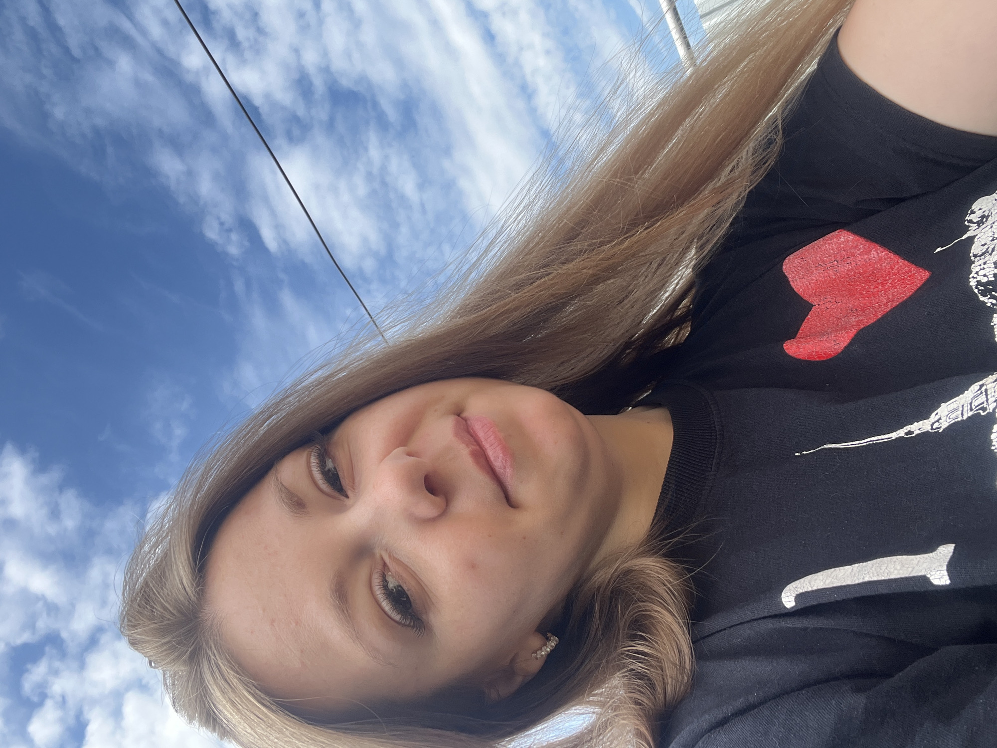 A young woman with blonde hair and blue eyes smiles at the camera in a close-up outdoor shot. She is wearing a black t-shirt with a graphic print that includes a red heart and a silhouette of the Eiffel Tower. The background is a bright blue sky with scattered white clouds. A thin wire stretches across the upper right portion of the image. The woman is wearing a small, sparkly earring in her left ear. The image appears to be taken in Shadrinsk, Russia, given the context.