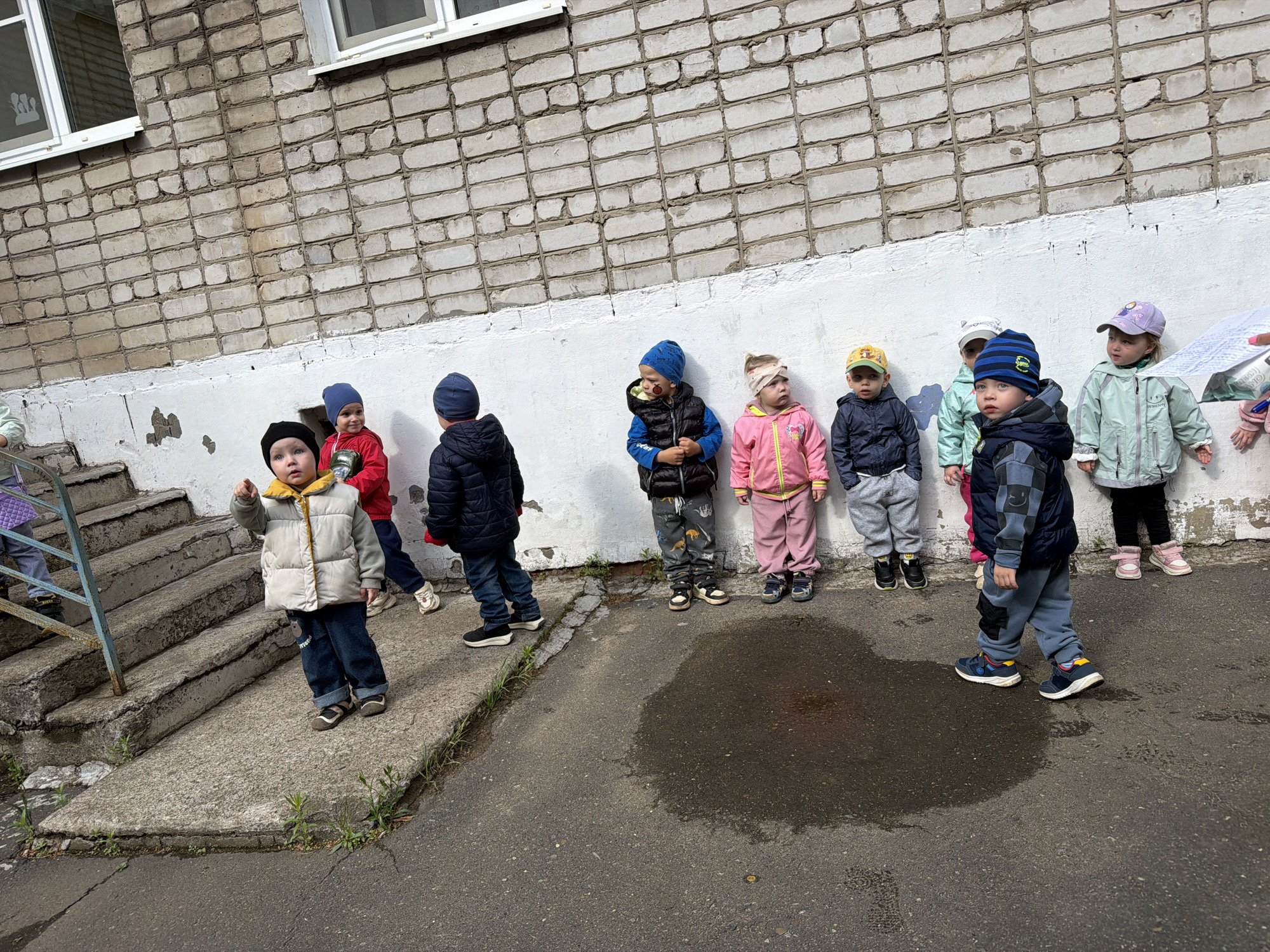 A group of approximately nine young children, likely toddlers or preschoolers, are gathered outdoors next to a weathered brick building with white window frames. They are dressed in warm jackets, hats, and long pants, indicating cool, overcast weather, further suggested by a prominent puddle on the asphalt ground in the foreground.
The children stand in a loose line against a white lower wall, which shows signs of wear and tear. On the far left, a child in a cream-colored puffer jacket points toward something out of frame. Another child in a red jacket to their right is smiling faintly. An adult's hand is visible on the far right, holding papers, possibly for an activity or instruction. The child in a light green jacket has the word "PRETTY" visible on her chest, and another child in a blue vest and dinosaur-patterned pants also appears to have "PRETTY" on his sleeve. A child wearing a green cap has "BUSH" visible on the front. On a window in the upper left, a white decal depicts a silhouette of three figures.
To the left of the children, concrete stairs with a metal railing lead downwards, also showing signs of age and scattered weeds. The overall scene suggests a supervised outdoor activity, possibly at a kindergarten or daycare in Chaikovsky, Russia. The children's expressions vary from curious to serious, reflecting a calm and organized atmosphere.