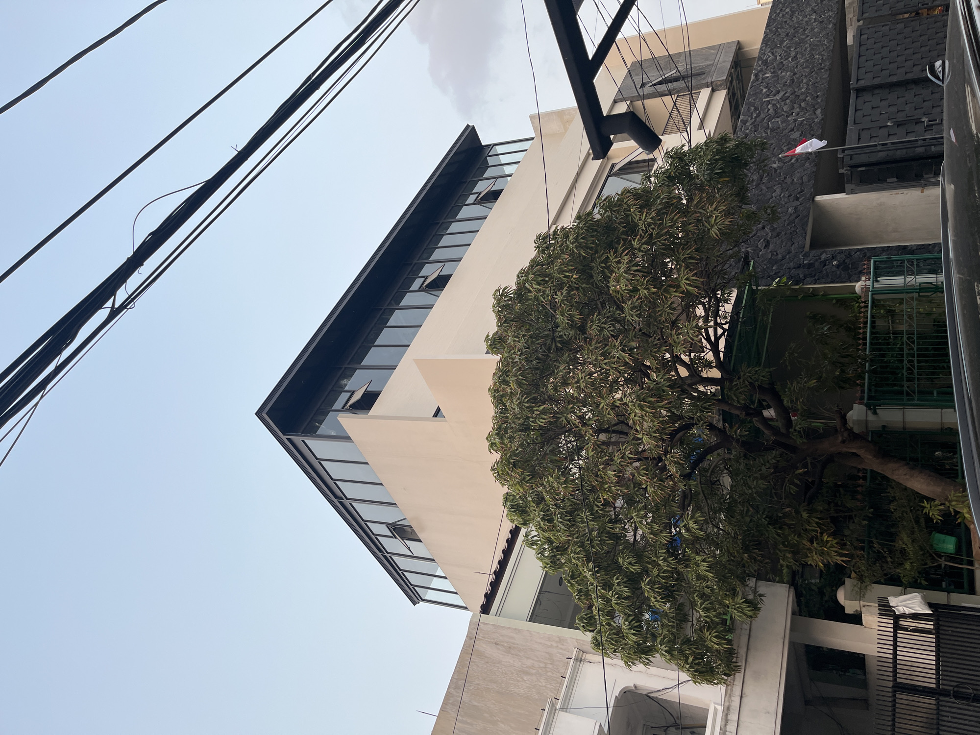 A low-angle, full-shot captures a building in an urban environment, with a clear sky overhead and a cluster of electrical wires diagonally spanning the upper portion of the frame. The building, painted a light cream color, features a modern upper story with a dark-framed, floor-to-ceiling glass facade. Several of the upper windows are slightly ajar. Below this, a section of the building is clad in dark, textured stonework, behind which an Indonesian flag is visible, partially obscured by a black metal fence. In the foreground, a mature tree with dense green foliage stands prominently, its branches reaching towards the building. To the left, the edge of another building with white architectural detailing is visible, along with a black metal gate. The lower part of the image shows the roof of a car.
