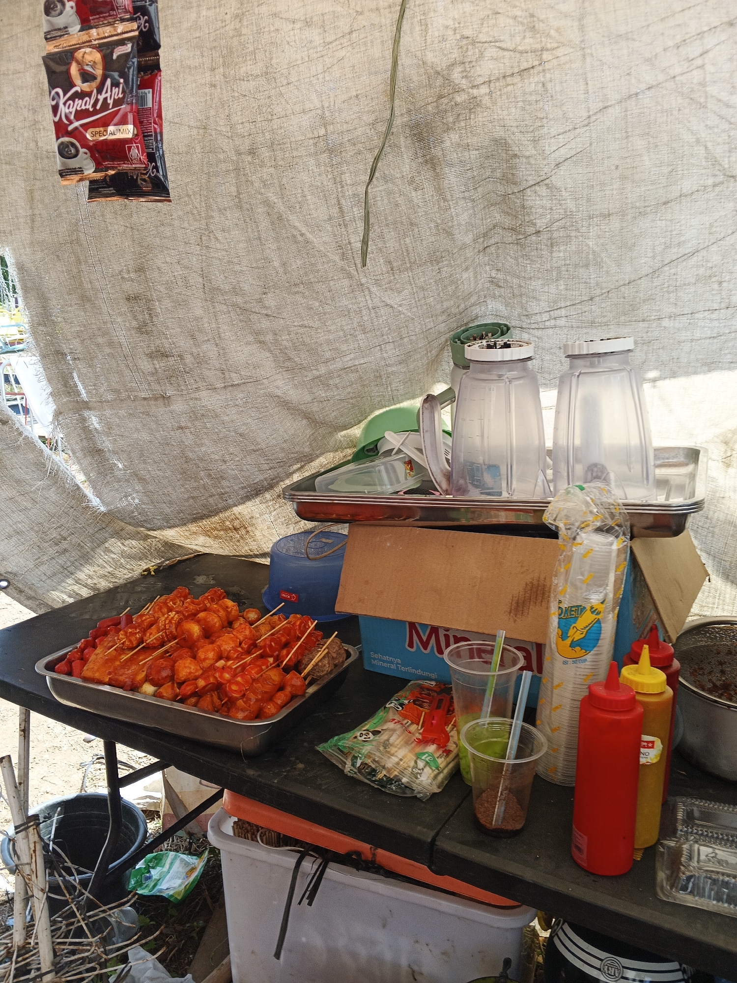 The image depicts an outdoor food stall, likely a temporary setup, in Sukabumi, Indonesia. A large, off-white tarpaulin is draped overhead, creating a covered area for the stall. Below this covering, a dark-colored table is laden with various food items and supplies.
In the foreground, a metal tray holds an assortment of skewers, featuring what appear to be grilled meat or seafood coated in a reddish sauce, along with some vegetable pieces. To the right of the tray, there are several plastic cups, some containing liquids and straws. Two condiment bottles, one red (likely ketchup) and one yellow (likely mustard), stand prominently, alongside a bag of plastic cups and a metal mesh container. Behind these, two clear blender pitchers are stacked on a silver tray, suggesting that drinks are also being prepared or served. A cardboard box, labeled with "Mino" and Indonesian text, is also visible.
In the background, hanging from the tarpaulin, are packets of "Kapal Api" brand coffee. The ground below the table is littered with debris, including plastic bags and some white piping, indicating a somewhat makeshift or informal setting. The overall scene suggests a bustling street food vendor preparing for or in the midst of serving customers. There are no people directly visible in the frame.