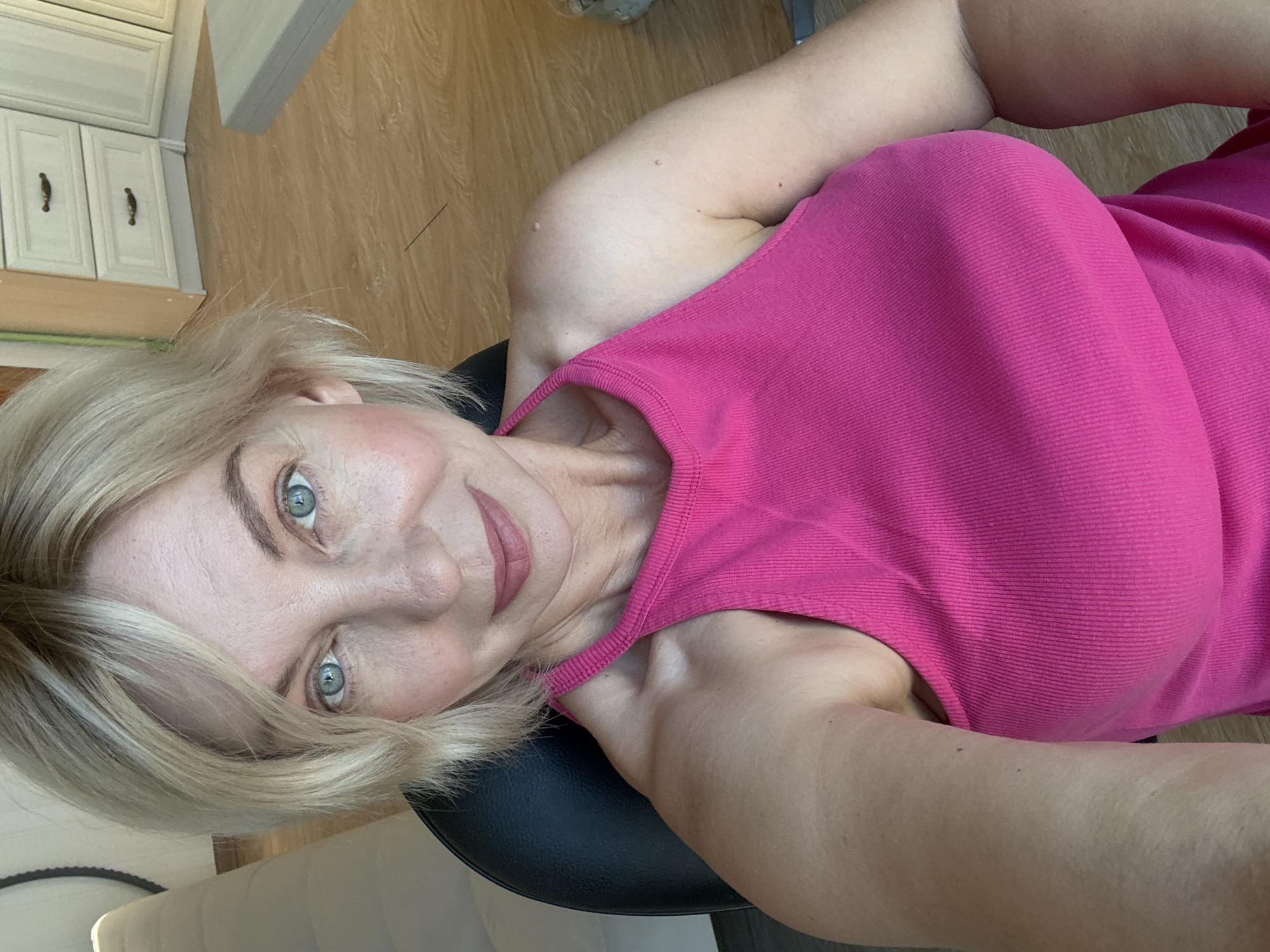 A woman with blonde hair and blue eyes, wearing a pink tank top, is seated in a black chair. She is looking towards the camera with a slight smile. The background shows a wooden floor and white cabinets.
