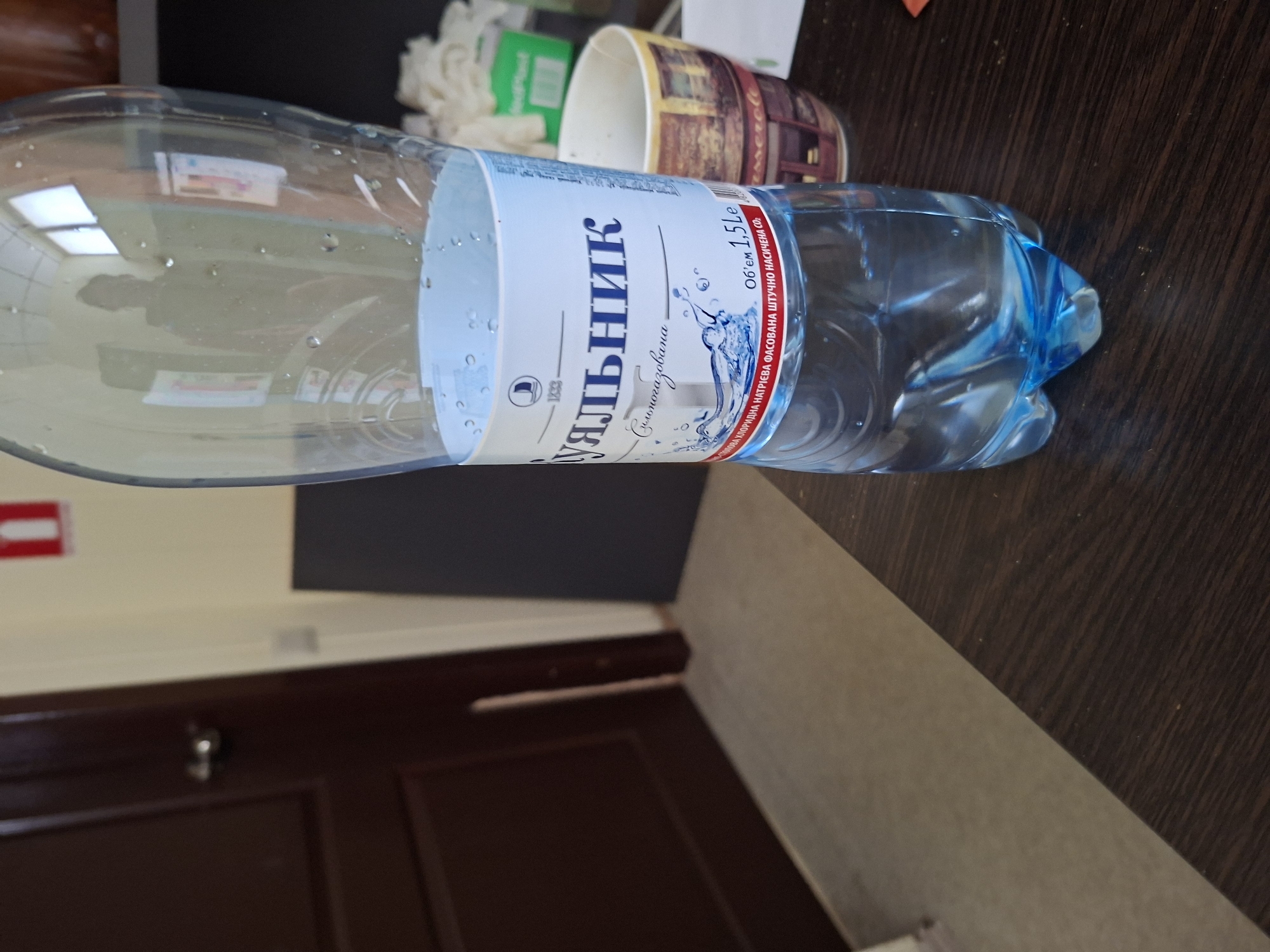 The image shows a plastic bottle of mineral water on a dark wooden table. The bottle has a blue and white label with Cyrillic text. To the right of the bottle is a patterned cup. In the background, there is a dark brown door with a silver doorknob and a portion of a wall. A red fire extinguisher symbol is visible on the wall. The scene appears to be indoors, possibly in an office or residential setting. The lighting suggests it is daytime. The image does not provide any specific information about the location in Kyiv, Ukraine, or any details about people or their actions.