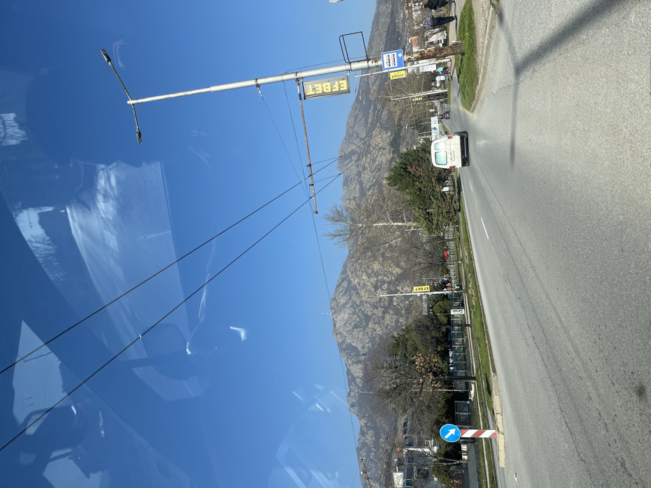 The image depicts a daytime scene on a multi-lane asphalt road in the city of Sliven, Bulgaria, with the Sinite Kamani mountain range prominently visible in the background under a clear blue sky. The perspective is from inside a vehicle, as indicated by reflections of a dashboard or windshield structure in the upper portion of the frame.
The road features a central grassy median containing trees, some bare and others evergreen. A blue circular traffic sign with a downward white arrow is mounted on a red and white striped post within this median, on the left side of the frame. On the right side of the road, multiple overhead wires span the scene, supported by metal utility poles. One such pole near the right edge displays a vertical yellow banner with the text "EFBET" and a blue rectangular bus stop sign. A white panel van is driving away from the camera in the right lane of the road. Several other vehicles are visible further ahead in the distance. On the sidewalk adjacent to the right lane, a group of approximately three to four pedestrians is walking away from the camera, near what appears to be a bus stop shelter. The mountainous background exhibits rocky terrain with sparse deciduous trees, suggesting a late winter or early spring season.