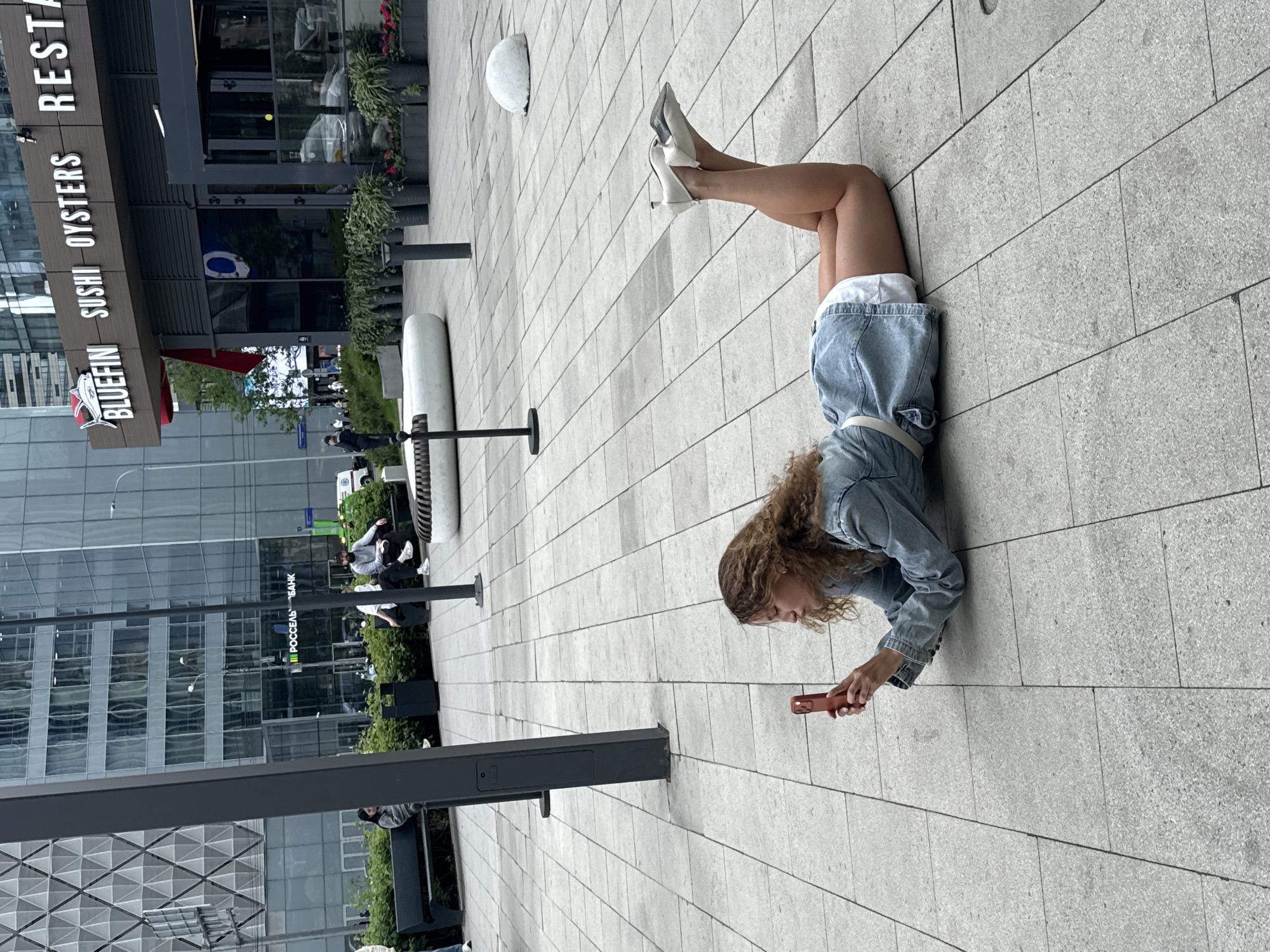 A woman lies on a gray, paved surface in Khimki, Russia, using a red mobile phone. She wears white high heels, a denim skirt, and a denim jacket. The pavement is patterned with rectangular slabs. In the background, buildings are visible, including one with the signage "BLUEFIN SUSHI OYSTERS RESTAURANT" and another with "РОССЕЛЬХОЗБАНК." There are benches, planters, and pedestrians in the distance.