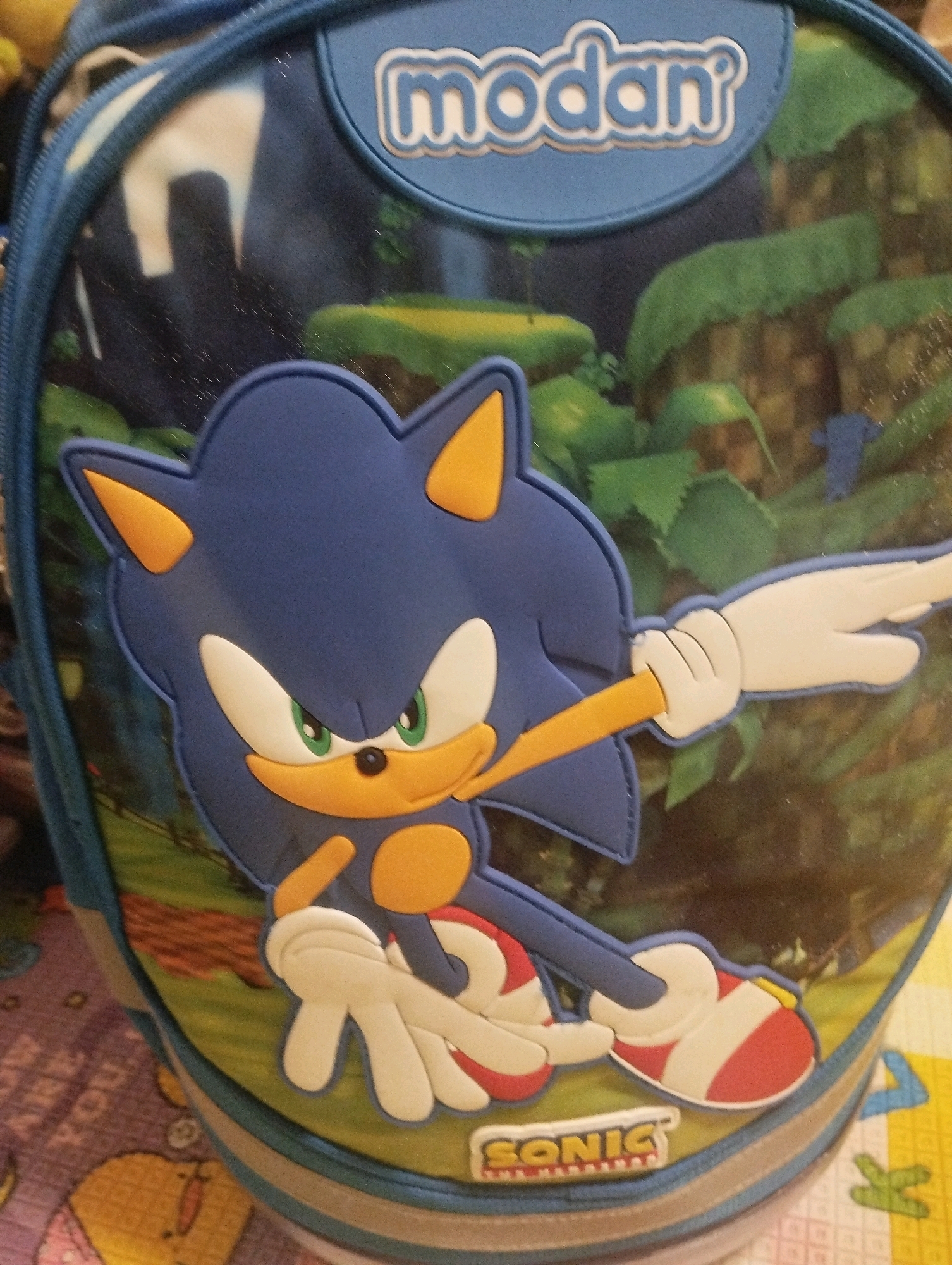This is a close-up, eye-level, indoor photograph of a blue backpack featuring a large, applique of the cartoon character Sonic the Hedgehog. The backpack has a blue fabric exterior with a lighter blue trim. A circular patch on the top of the backpack displays the word "modan" in white lettering with a light blue outline. The Sonic applique is prominent, showing the character's blue head and body, his signature green eyes, yellow muzzle and ears, and white gloves with outstretched fingers. He is depicted in motion, with his body angled and his legs bent, wearing red and white sneakers. The background of the backpack features a blurred image of a green, jungle-like environment with some indistinct white shapes. The backpack is resting on a surface that appears to be a patterned mat with yellow, pink, and purple sections, and some faint text is visible on the mat. The lighting suggests an indoor setting, possibly a room. The photograph does not depict any specific city or location.