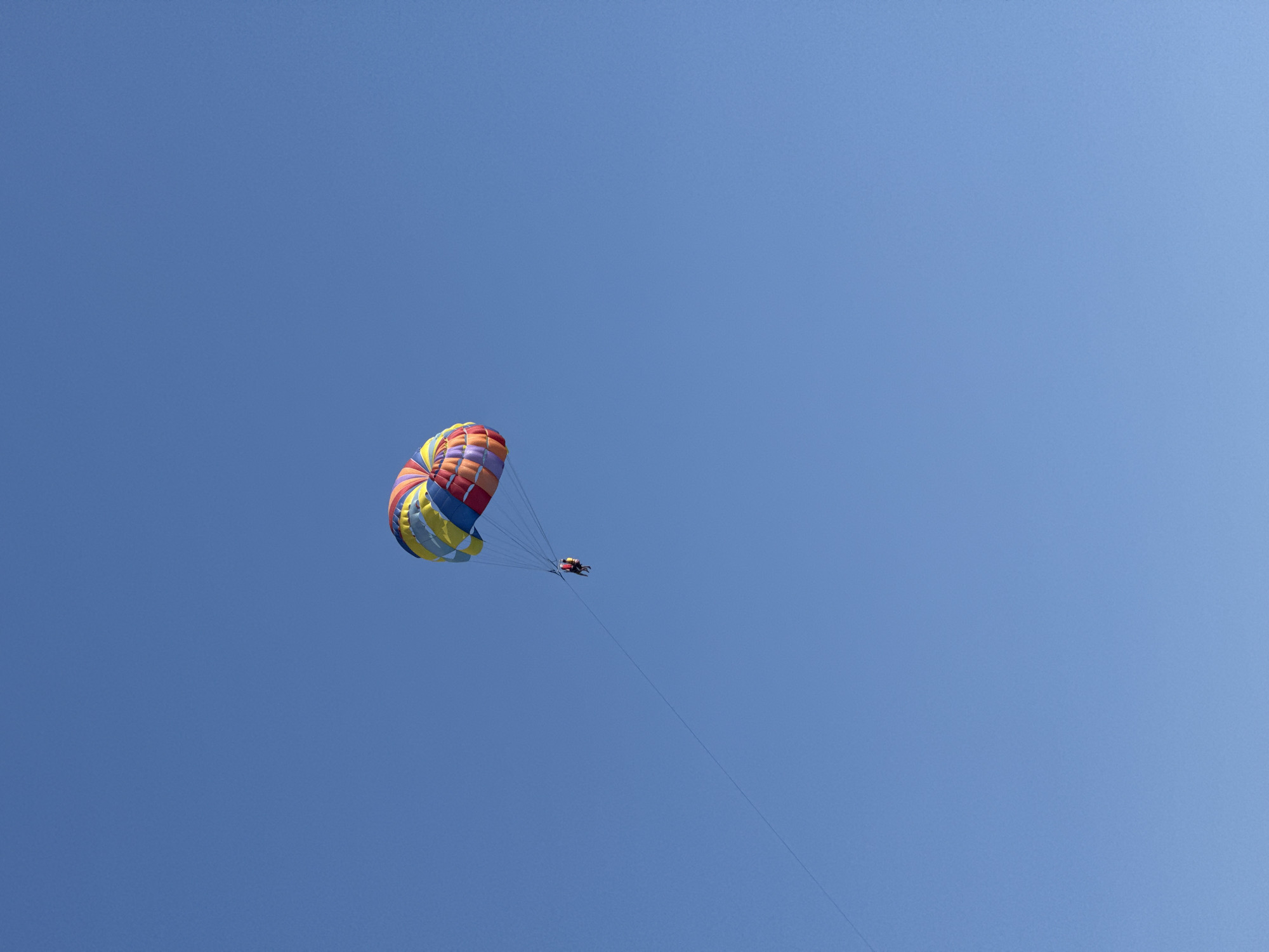 The image shows a parasailer suspended in a clear blue sky. The parasail is colorful, with segments of red, yellow, blue, and white. Two people are visible in the harness attached to the parasail, appearing to be enjoying the activity. A long tow rope extends diagonally downward from the parasail, disappearing out of frame. The scene is set against a vast expanse of unbroken blue sky. The location is identified as Kamenovo, Montenegro.