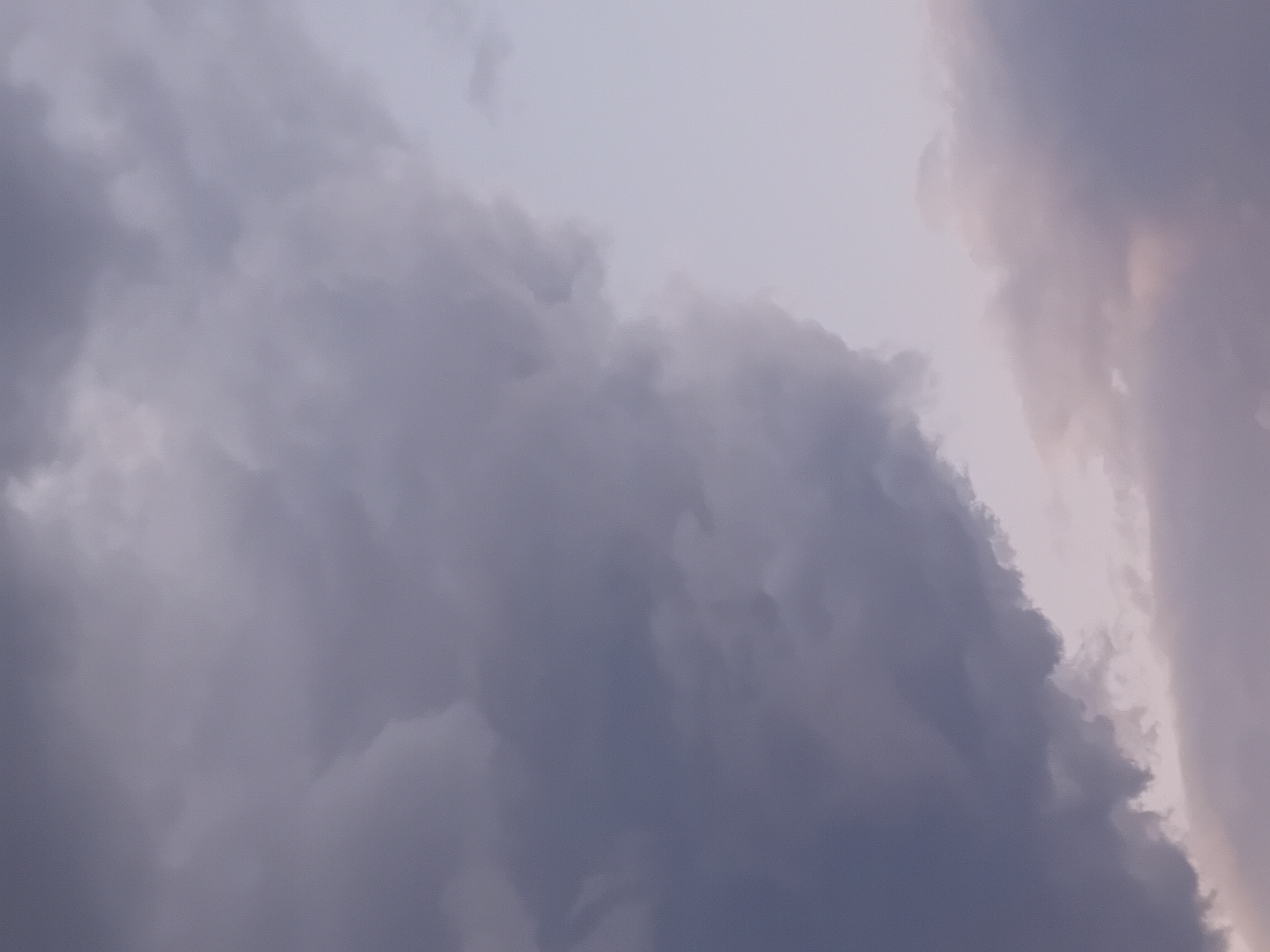 This is a close-up, low-angle photograph of a sky filled with dramatic, dark storm clouds. The clouds are predominantly shades of gray and purple, with some lighter, softer clouds visible towards the bottom and right of the frame. The lighting suggests it is either dusk or dawn, or that the storm is obscuring the sun. There are no people, buildings, or landforms visible, indicating the photograph was taken from an elevated position or focused solely on the sky. The scene is forensically devoid of any specific identifying landmarks within the city of Onikişubat, Turkey.