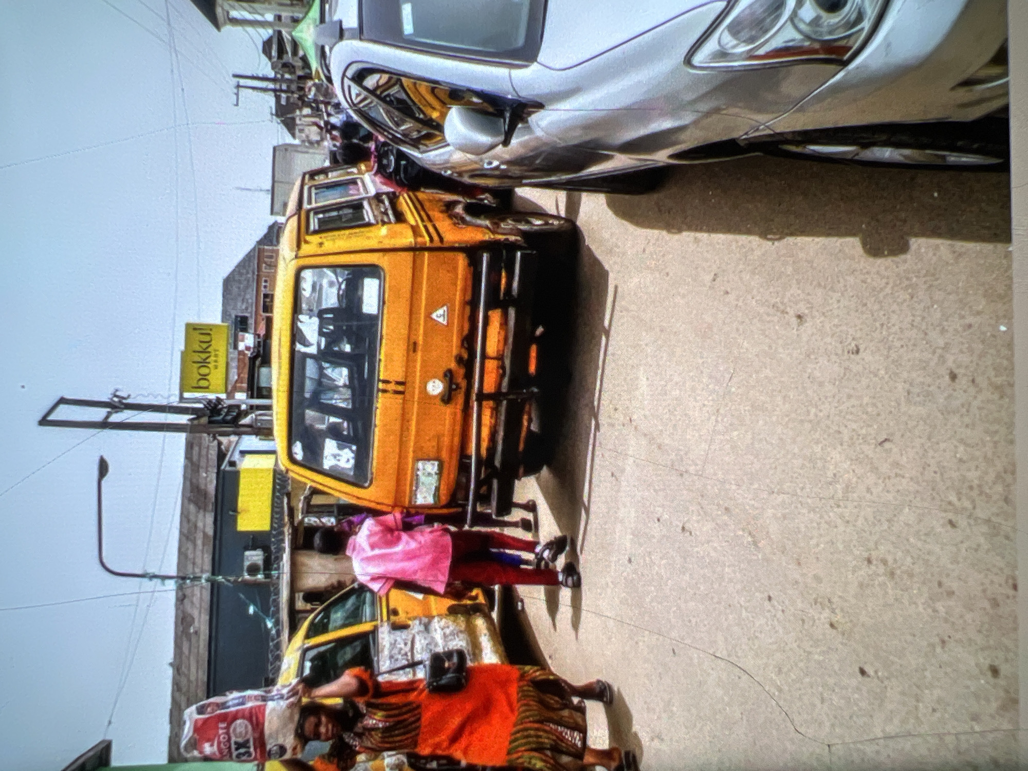 In Lagos, Nigeria, a street scene unfolds with a palpable sense of daily life. The foreground is dominated by the parked silver sedan, its sleek lines contrasting with the rougher textures of the street. To its left, an orange public transport van, a common sight in Lagos, is positioned. The van, with its characteristic yellow and black livery, appears to be waiting or perhaps stuck in traffic. A woman in a vibrant orange and patterned dress walks away from the viewer, carrying a large white bag labeled "ANGOTE 3X" on her head. She is followed by a young man in a pink shirt and dark pants, also walking away from the camera. The background reveals a bustling urban environment with buildings, power lines, and streetlights, indicative of a busy commercial area. A yellow sign with "bokku!" in bold letters hangs above a storefront, suggesting a commercial establishment. The overall impression is one of a typical day in Lagos, where commerce, transportation, and pedestrian activity converge.