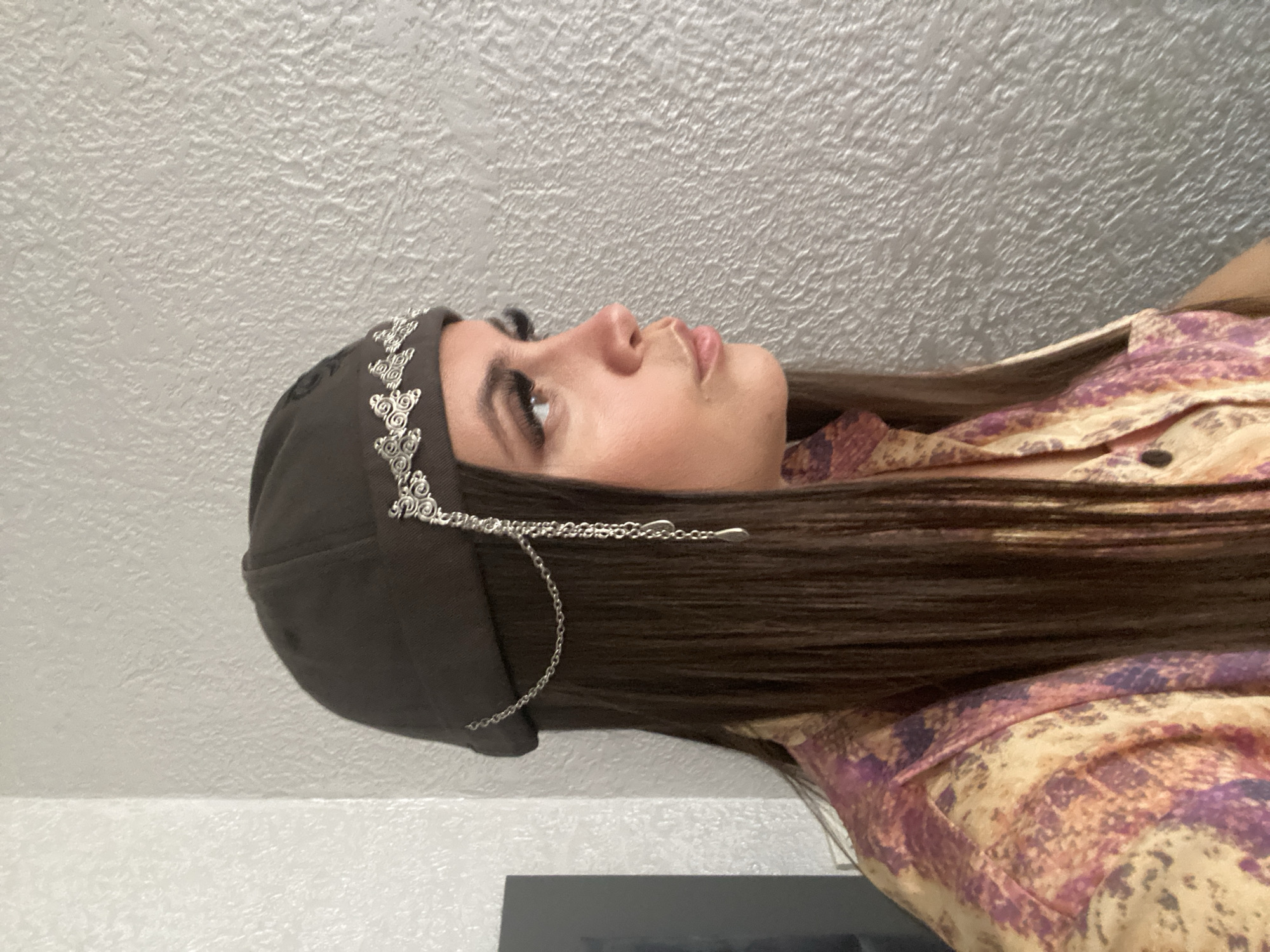 A woman is shown in profile, facing right, against a textured light grey wall. She is wearing a dark brown cap adorned with a silver filigree band and a dangling silver chain with a teardrop pendant. Her long, dark brown hair cascades down her shoulders, partially covering a colorful shirt with a pattern of abstract shapes in shades of yellow, orange, and purple. Her makeup includes prominent false eyelashes and defined eyebrows. The lighting suggests an indoor setting. The location is not identifiable as Chalda, Russia.