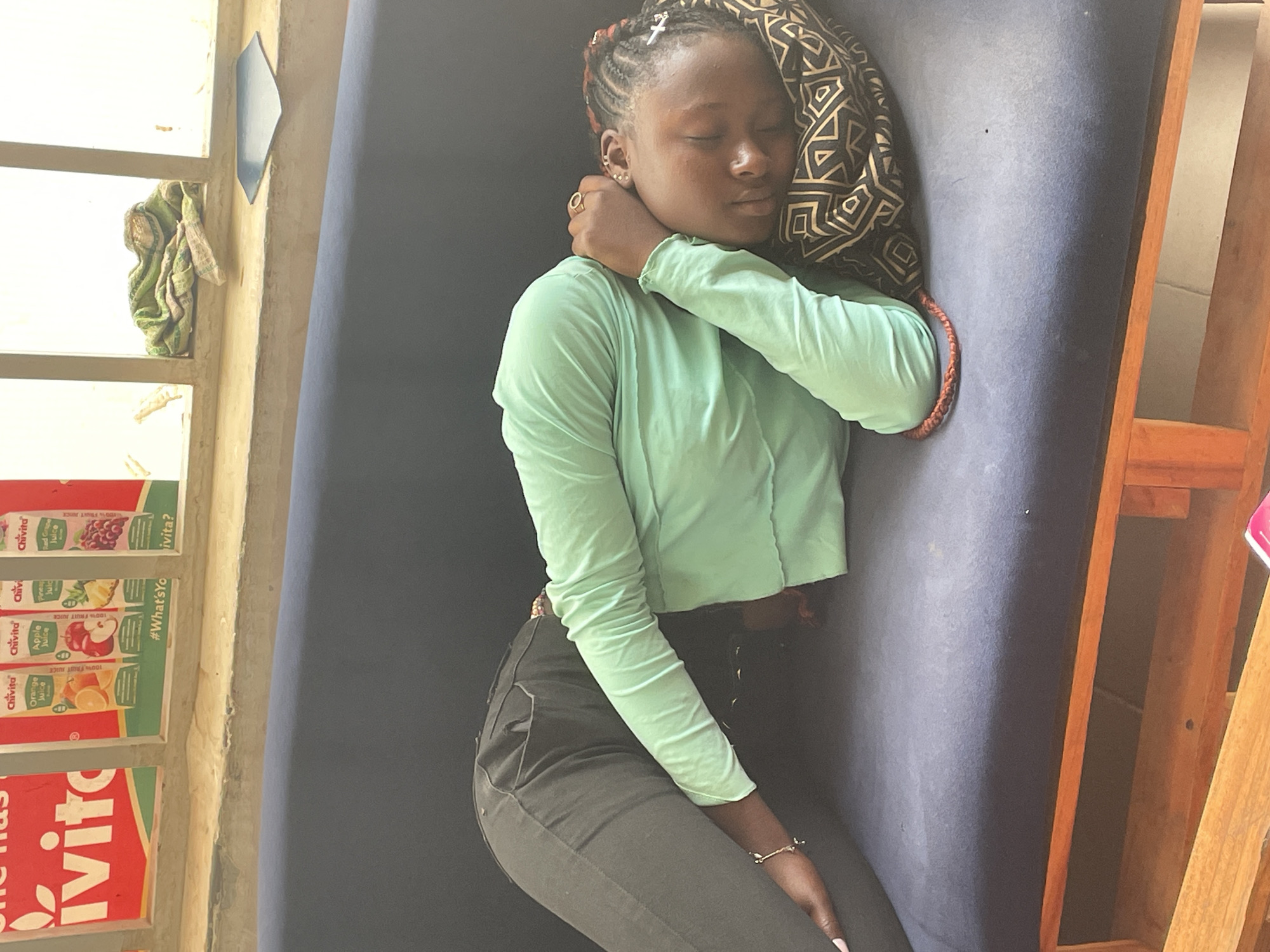 A young woman is depicted resting or sleeping indoors, positioned on her side on a dark blue mattress or couch. Her eyes are closed, conveying a sense of peace or deep rest. She has intricately braided hair adorned with small white accessories and partially covered by a black and gold patterned headscarf or fabric. She wears a light mint-green long-sleeved top and dark trousers. A gold-toned ring is visible on her finger, and a delicate bracelet graces her wrist.
To her left, a window provides bright illumination, suggesting daytime. A light green and white patterned cloth is tucked into the wooden window frame. Below the window, a stack of Chivita juice boxes is prominently displayed, showing various flavors such as Red Grape, Pineapple, Apple, and Orange. Visible text on the boxes includes "Chivita", "100% FRUIT JUICE", and "#What'sYo ivita?". To her right, a simple wooden frame, possibly part of a bed or furniture, is visible. The presence of the Chivita brand is a strong indicator for a Nigerian setting, consistent with the provided location context of Barakin Akawo, Nigeria.
