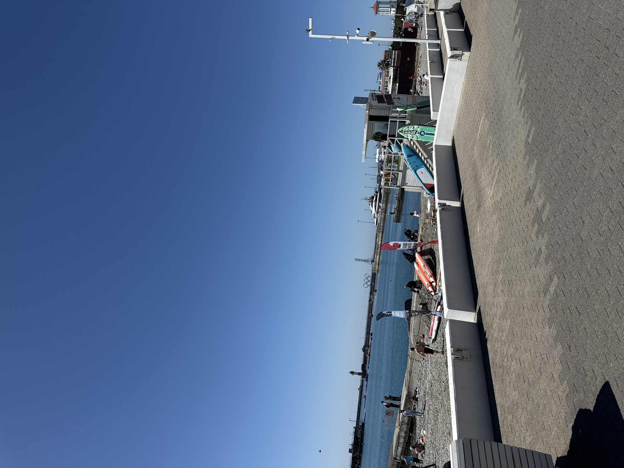 The image depicts a vibrant waterfront scene under a clear blue sky. The perspective is from a raised paved walkway or promenade, which occupies the right side of the frame and features a patterned grey surface. A white railing separates this elevated path from a lower area where various water sports equipment is stored or displayed.
In this lower section, numerous stand-up paddleboards are visible, including a prominent green one marked with the number "5" and a blue one. Several orange and black/white sails or kites, likely for windsurfing or kiteboarding, are also propped up. Beyond this equipment, a pebbly beach stretches to the blue water.
Numerous people are scattered along the beach and at the water's edge, some appearing to relax, others potentially engaging in or preparing for water activities. A red circular sign with a diagonal line, indicating a prohibition, is visible near the water.
Across the expanse of blue water, a distant lighthouse stands tall. Further along the opposite shore, a large structure resembling the Olympic rings is clearly visible, hinting at a location like Sochi. Various buildings, including white structures and one with a red-tiled roof, line the distant coastline. The bright, cloudless sky and distinct shadows suggest it is a sunny day, likely morning or early afternoon. The overall scene portrays an active recreational area or marina.