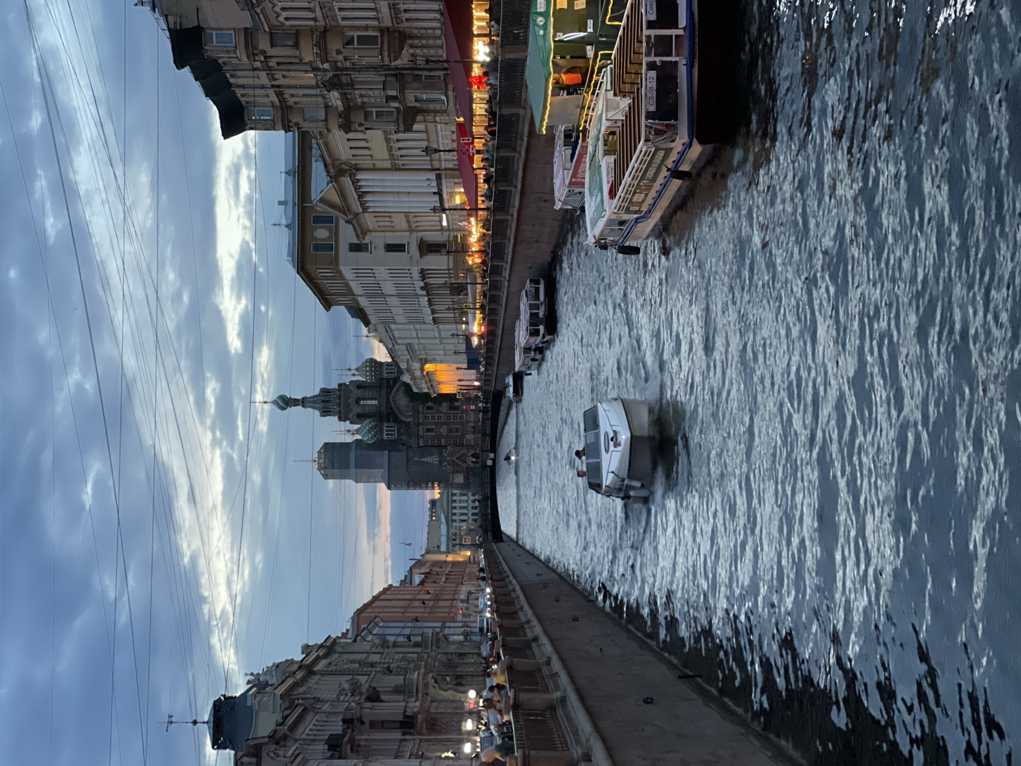 The image depicts a canal in Saint Petersburg, Russia, likely the Griboyedov Canal, given the presence of the Church of the Savior on Spilled Blood in the background. It is dusk, with a cloudy sky overhead and lights illuminating the buildings lining the canal. Several boats are present on the water, including a small white speedboat in the foreground with two individuals on board, and several larger tour boats docked or moving along the canal. People are visible on the sidewalks along the canal, some appearing to be dining or walking. The architecture is characteristic of Saint Petersburg, with ornate building facades. The scene conveys a sense of urban life and tourism in the early evening.