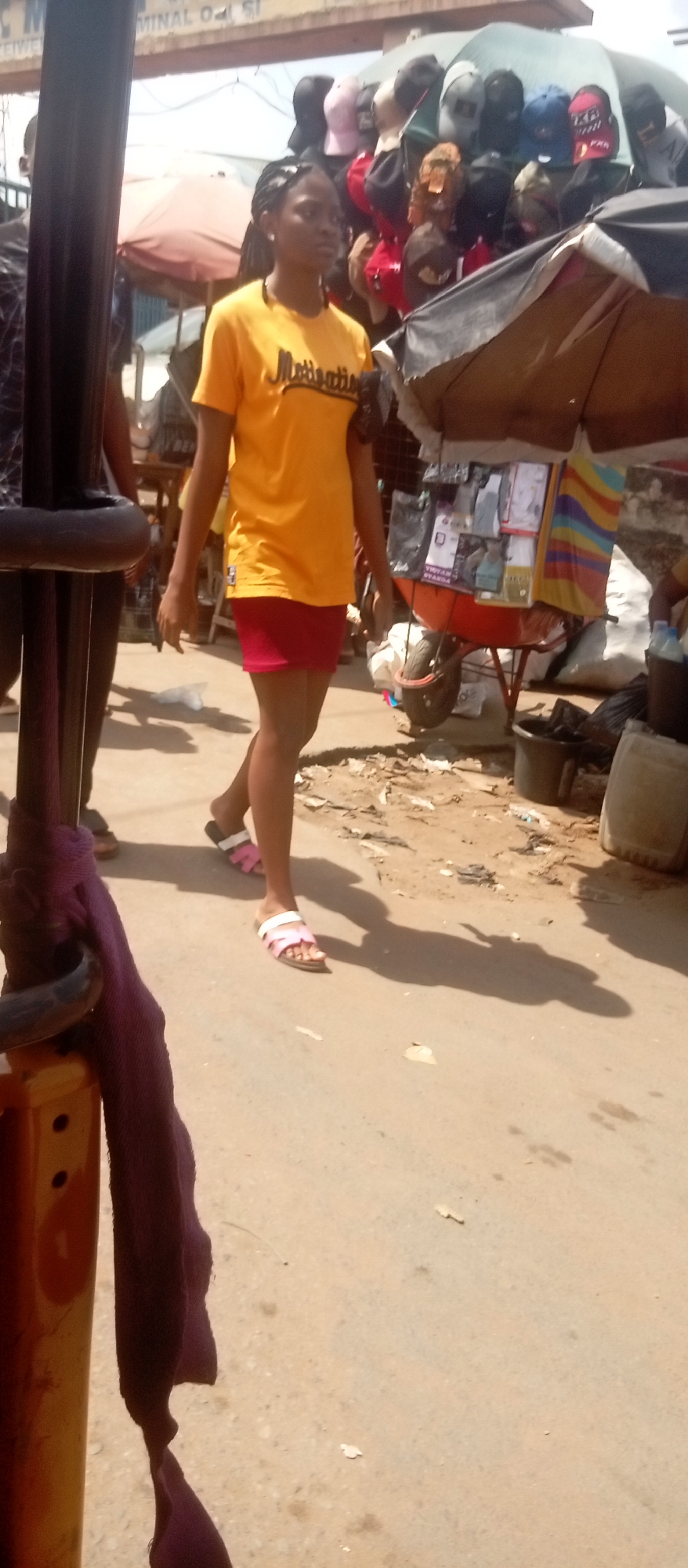 On a sunny day in Onitsha, Nigeria, a young woman walks along a street market. She wears a yellow t-shirt with "Motivation" printed on it, a red mini skirt, and pink sandals. Behind her, vendors sell merchandise, including baseball caps displayed under an umbrella. A man walks on the left side of the frame. The foreground includes part of a yellow and black auto-rickshaw with a piece of purple cloth draped on it. The ground is paved with dirt and debris.