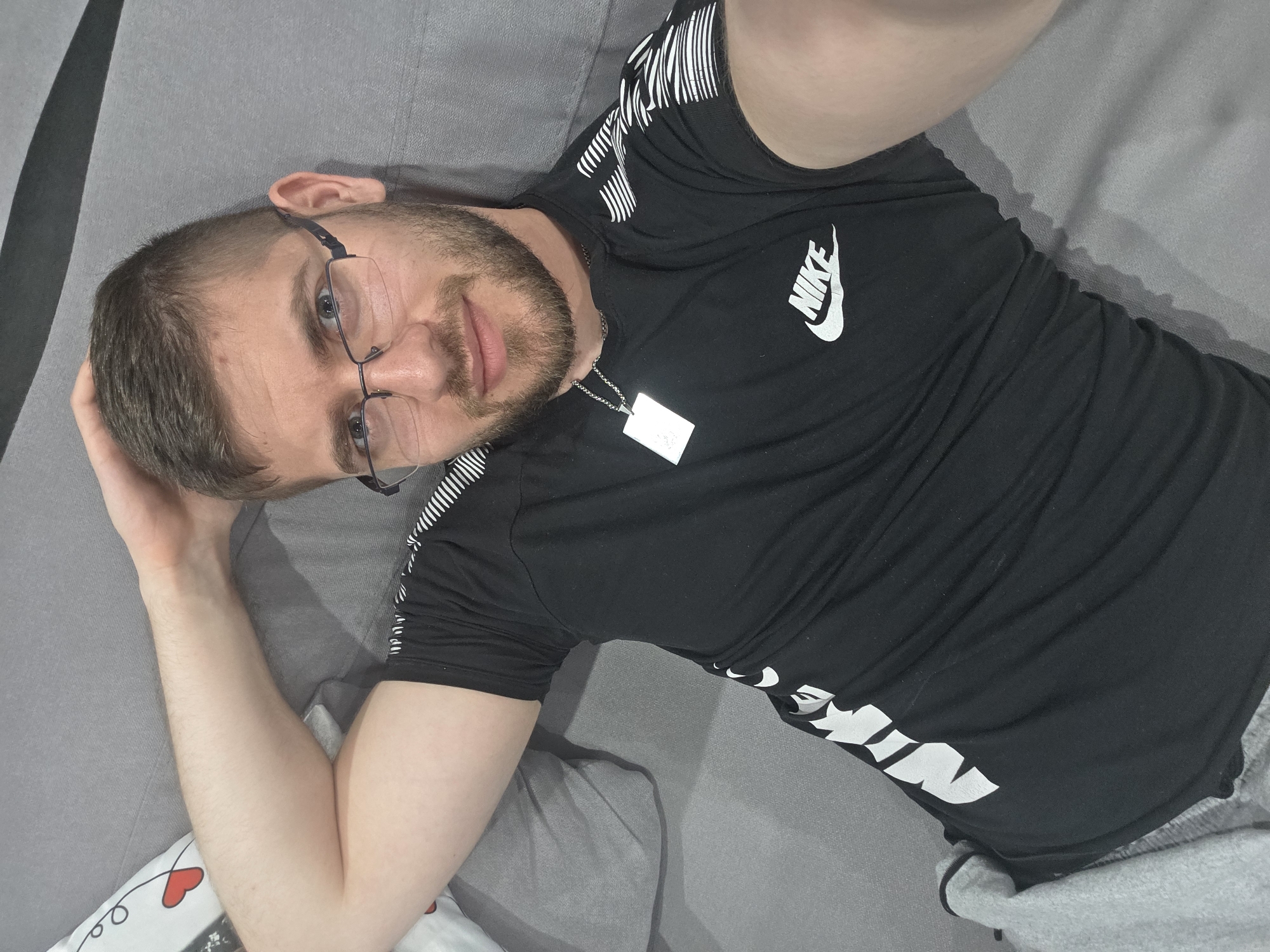 An adult male is captured in an indoor setting, lying on a grey sofa or bed in Yagodovo, Bulgaria. He has short brown hair, a neatly trimmed beard, and wears dark-framed eyeglasses. He is dressed in a black t-shirt with a prominent white "NIKE" logo on the chest, complemented by white stripe patterns on the shoulders and sleeves. A silver chain necklace with a rectangular pendant hangs around his neck. Grey sweatpants are partially visible on his lower body.
The man is lying on his back, with his head slightly turned to face the camera. His left arm is positioned behind his head, while his right arm rests by his side. He looks directly at the viewer with a relaxed expression and a slight smile. The background primarily consists of the plain grey upholstery of the furniture. A white pillow with a red heart design is visible in the bottom left corner of the frame. The lighting suggests it is daytime indoors.
