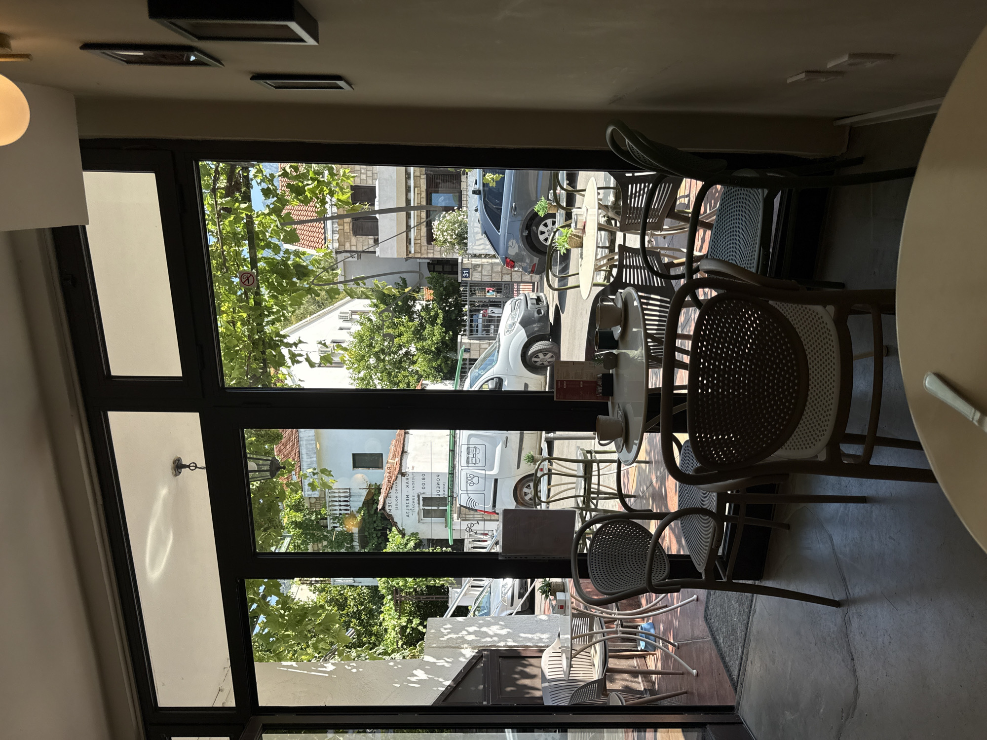 A cafe with large glass windows overlooks a street in Budva, Montenegro. Inside, several tables and chairs are arranged, some with cups of coffee. Outside, parked cars and buildings are visible. The sun casts shadows through the foliage.