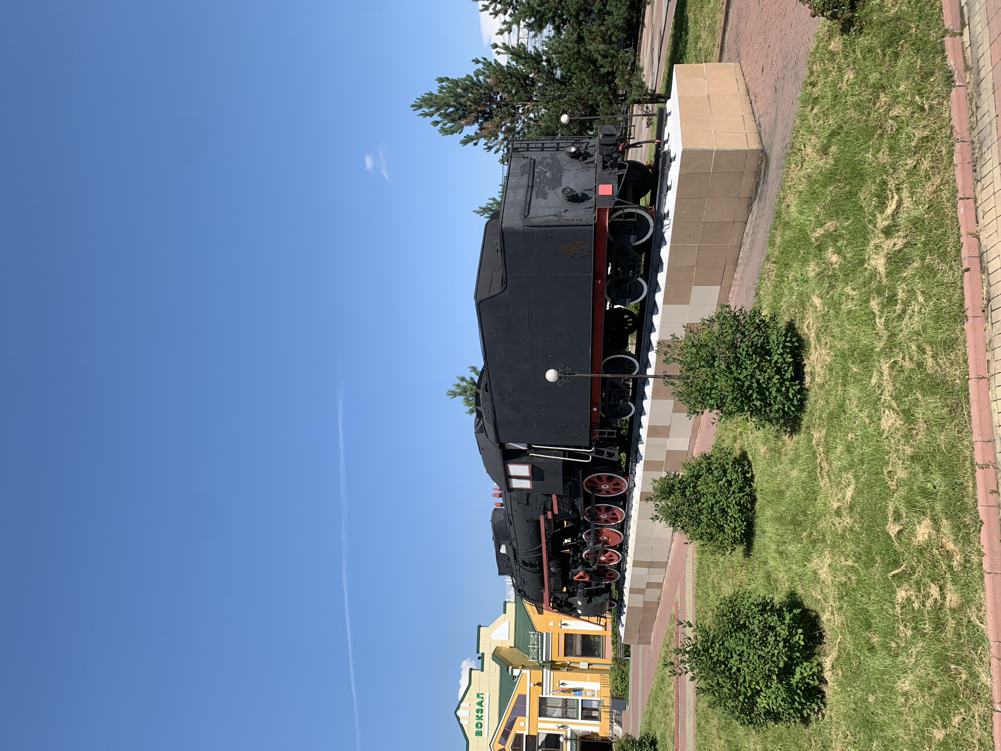 The image shows a preserved steam locomotive on display outdoors in front of a train station building. The locomotive is black with red-rimmed wheels and is mounted on a pedestal designed to look like train tracks. The train station building is yellow and green and has the word "ВОКЗАЛ" (VOKZAL, meaning "train station" in Russian) written in large green letters on its facade. Several green bushes are visible in the foreground, and pine trees are to the right of the locomotive. The sky is clear and blue with a faint contrail. This scene is located at the Prokopyevsk railway station in Prokopyevsk, Russia. There are no people visible in the image.