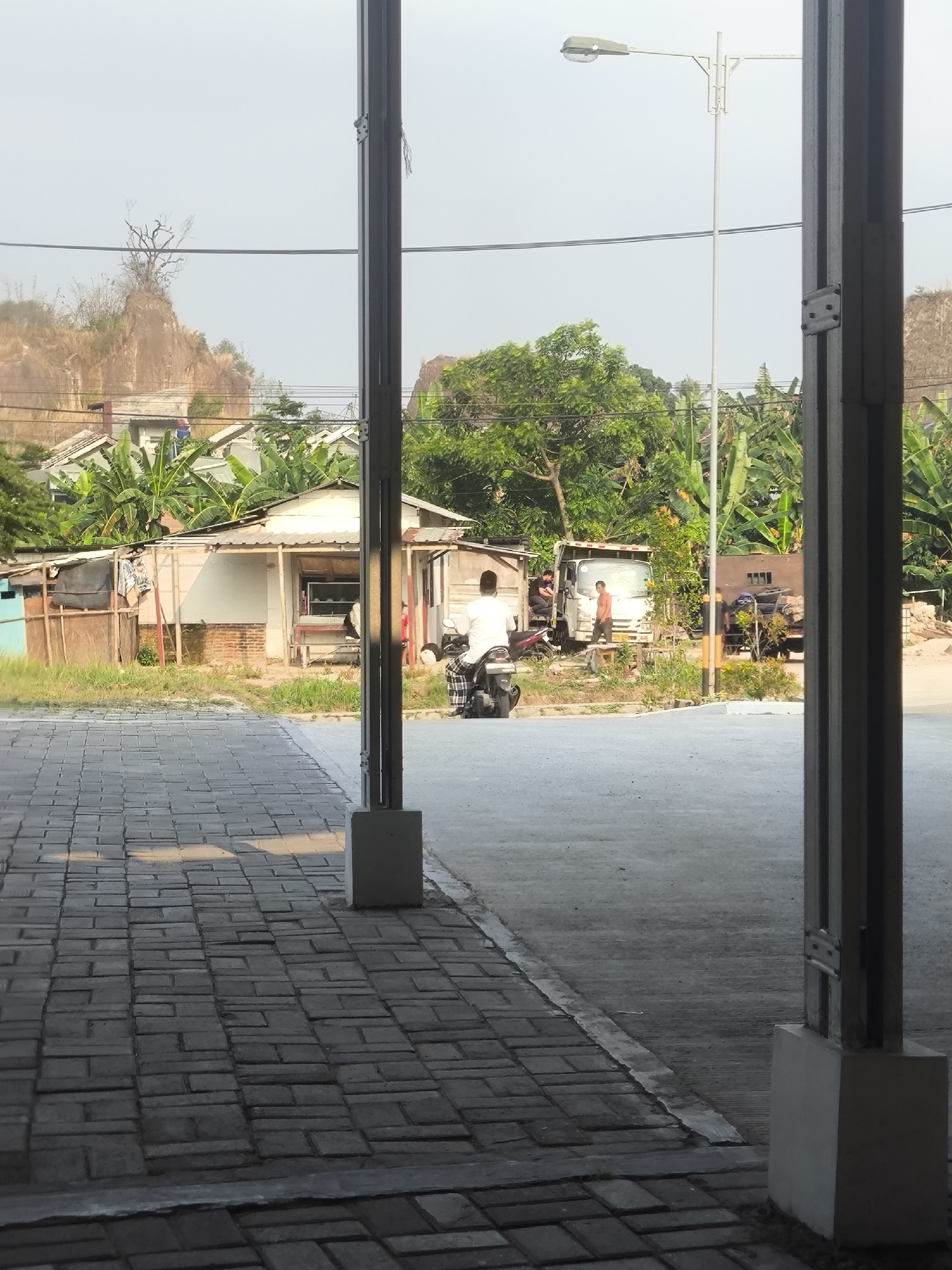 The image depicts an outdoor scene with a paved walkway in the foreground, transitioning to a dirt and grass area. A person wearing a white shirt and patterned pants is seated on a motorcycle, facing away from the camera. In the middle ground, there is a small, simple building with a white roof, possibly a shop or house, with a red bench in front. Adjacent to this structure, a white truck is parked, and several individuals are visible near it. Further in the background, lush green trees and banana plants are abundant, suggesting a tropical environment. A rocky hill with sparse vegetation rises behind the buildings. A street light stands tall to the right of the frame. The scene appears to be in Cilegon, Indonesia, given the presence of banana trees, tropical vegetation, and common building styles. The overall atmosphere is that of a typical day in a semi-urban or rural area of Indonesia.