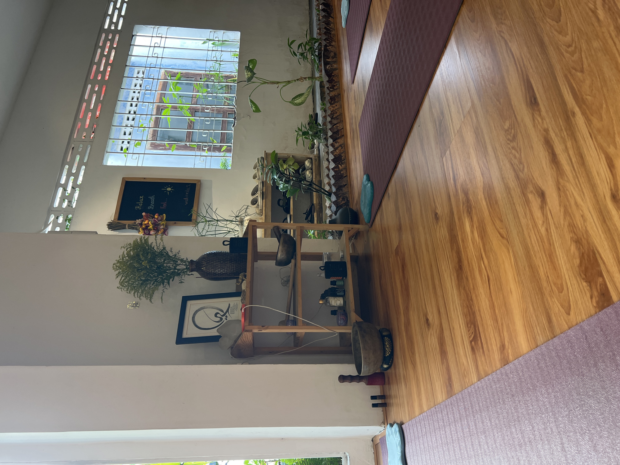 This is an interior shot of what appears to be a yoga studio or wellness space, likely in Nha Trang, Vietnam, given the architecture and plants visible through the window. The room is predominantly decorated in muted earth tones and features a light-colored wooden floor. Yoga mats, one light purple and another dark red, lie on the floor.
A wooden shelving unit is against the wall, filled with items such as shoes, decorative plants, singing bowls, and assorted bottles. On the wall above the shelving unit hangs a framed Arabic calligraphy artwork, as well as a chalkboard with a sun drawing and the words "Relax, Breath, Feel..." The room is brightly lit by natural light streaming through a window fitted with a security grill. Outside the window, green foliage is visible, suggesting a tropical or subtropical climate. The photograph is rotated clockwise.