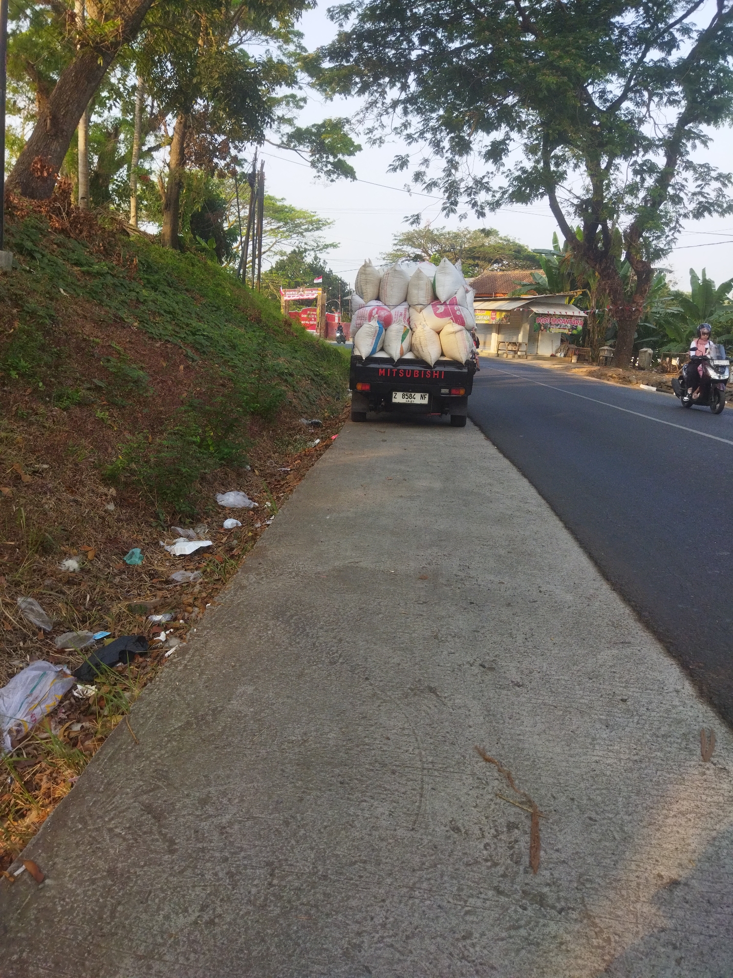 The image depicts a roadside scene in Banjar, Indonesia. A black Mitsubishi pickup truck is parked on the side of a paved road, heavily loaded with large sacks, likely containing agricultural products. To the left of the truck, there is a dirt embankment covered with vegetation and scattered litter, including plastic bags and leaves. The road itself is asphalt, and to the right, a person on a scooter is visible. In the background, there are trees, buildings, and some signage, including what appears to be a local shop. The overall impression is one of a rural or semi-urban setting with signs of commercial activity and some environmental neglect.