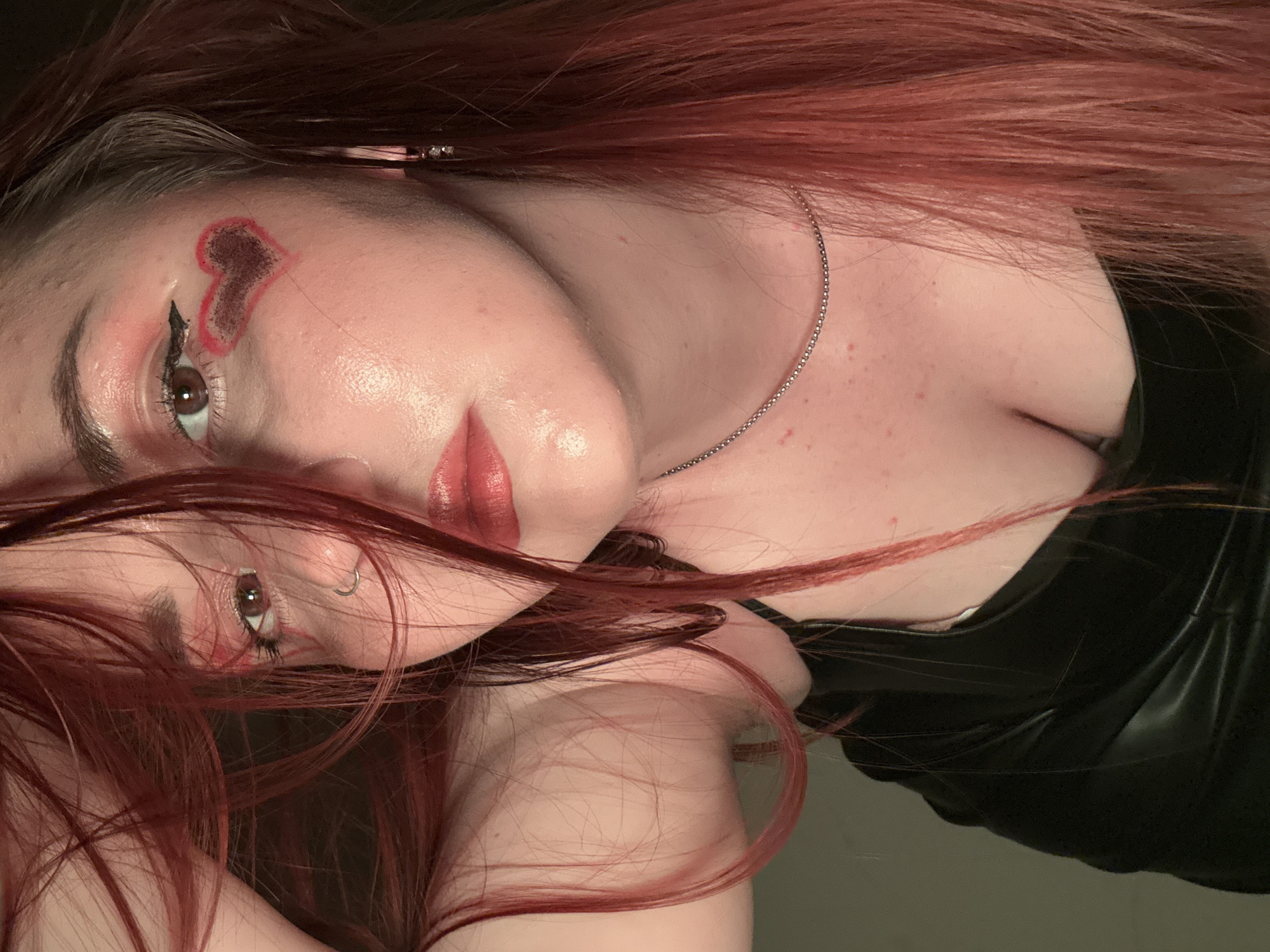 A woman with red hair is pictured close-up. She is wearing makeup including a heart on her forehead and red lipstick. She has a nose ring and is wearing a silver chain necklace. She is wearing a black garment. The image is rotated. There is no indication of the location being Izhevsk, Russia.
