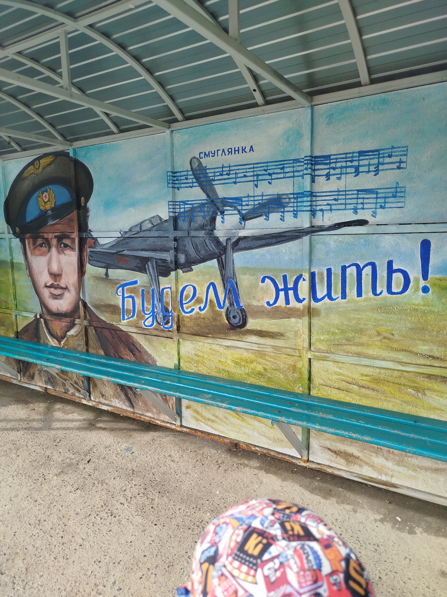 This media file depicts a mural painted on the exterior wall of a bus stop shelter in Zavodoukovsk, Russia. The mural features a portrait of a man in a pilot's uniform, including a cap with a red star emblem, alongside a propeller-driven airplane. Musical notes are depicted in the sky behind the airplane, and the text "Смуглянка" is visible above them. Below the airplane, the Cyrillic words "Будем жить!" are painted in large, blue letters, which translates to "We will live!". The style of the painting is somewhat rough and appears to be hand-done.
The scene is set outdoors, under the roof of the bus stop shelter, which has a corrugated metal roof supported by white metal beams. A turquoise metal bench is positioned in front of the mural. The ground is paved with concrete, and there is evidence of rust on the metal structure supporting the bench. The background of the mural depicts a yellow and green landscape, possibly representing a field, under a light blue sky. The overall mood evoked by the mural, with the pilot, the airplane, and the defiant phrase "We will live!", suggests a theme of resilience, possibly related to wartime or post-war sentiment. The presence of the bench and the shelter indicates it's a public space designed for waiting. No specific time of day or weather conditions are definitively indicated, though the lighting suggests daytime.