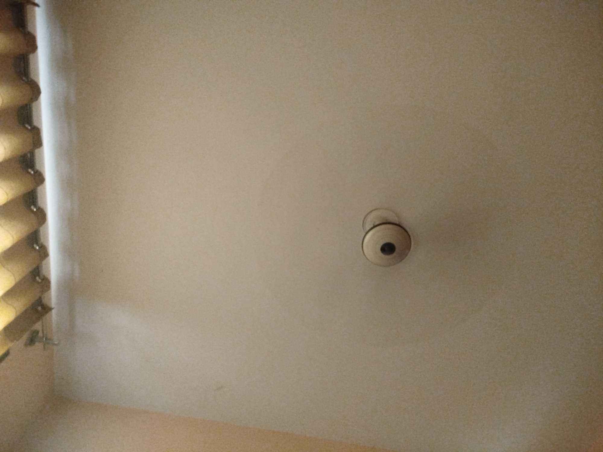 The image shows a ceiling with a light fixture and blinds on the left side. The scene is indoors, likely in a room. There are no people or animals visible in the image. The ceiling is a light beige color, and the blinds are a golden yellow. The light fixture appears to be a simple dome-shaped fixture with a circular grille. The overall lighting in the room is dim, with a shadow cast by the blinds on the wall. There is no information in the image to suggest the location is in Lassan, Pakistan.