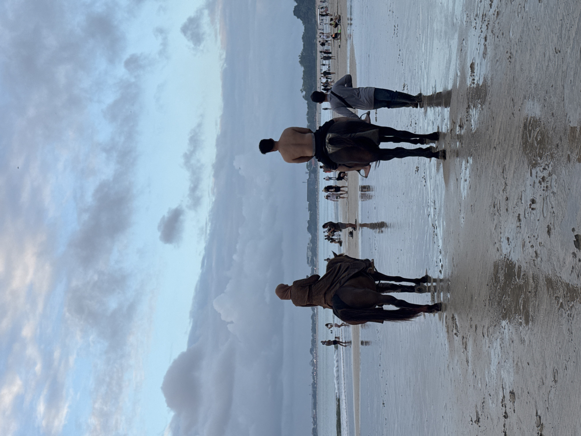 Two people are riding horses on a beach in Kuta Selatan, Indonesia. The person on the left is wearing a brown headscarf and a long brown dress. The person on the right is shirtless and wearing grey pants. They are both facing away from the camera. A third person, a male, is walking behind the horse on the right, wearing a grey shirt and blue jeans. There are other people in the background, as well as waves crashing on the shore and buildings in the distance. The sky is cloudy and the lighting suggests it is either dawn or dusk. The sand is wet and reflective, with footprints visible.