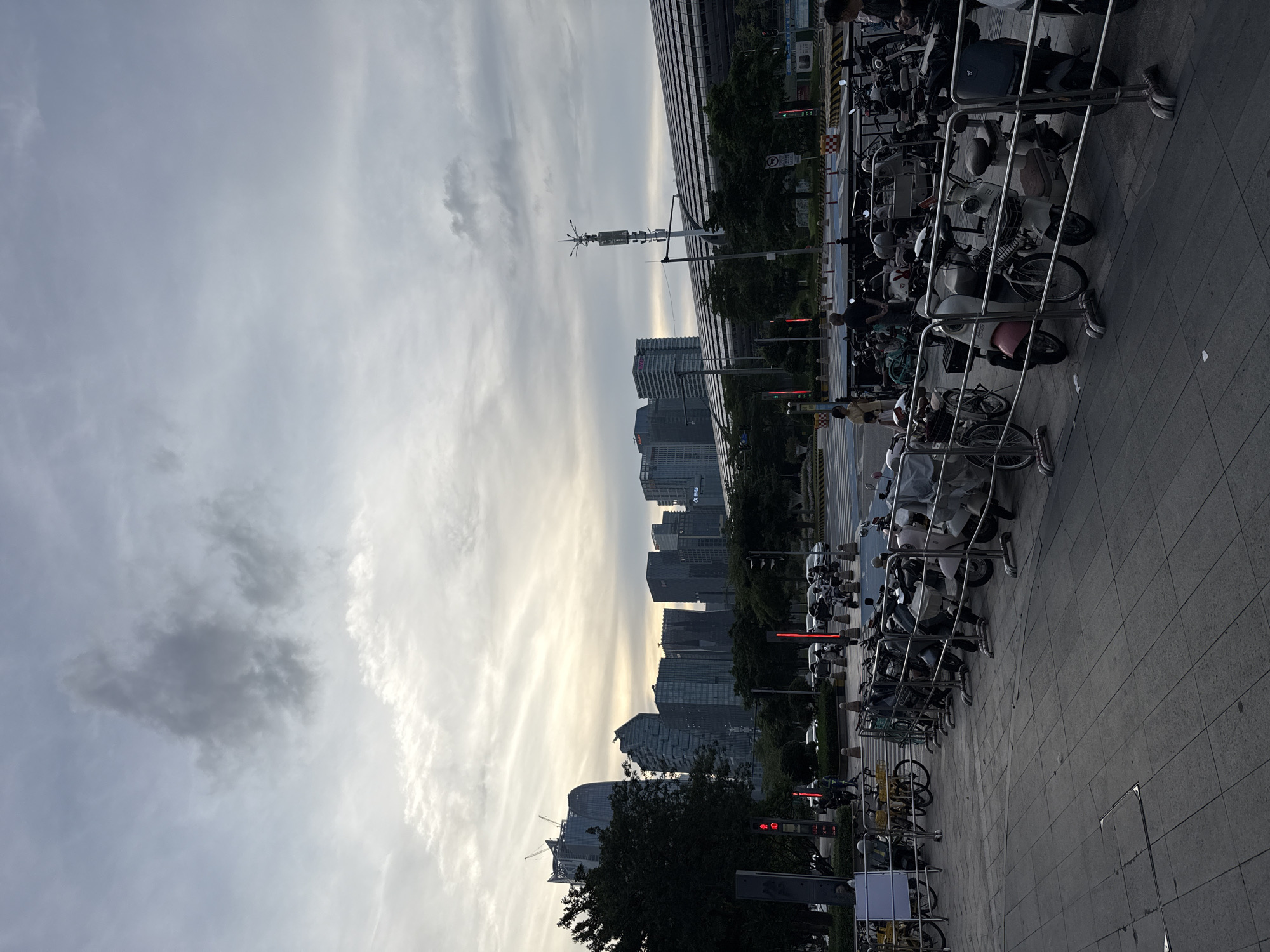 The image captures a street scene in Haizhu District, China. A row of parked bicycles and motorbikes are secured along a sidewalk in the foreground. Behind the sidewalk are trees, street lights, and several modern high-rise buildings. The sky is overcast with grey and white clouds. In the far distance, a radio tower is visible.