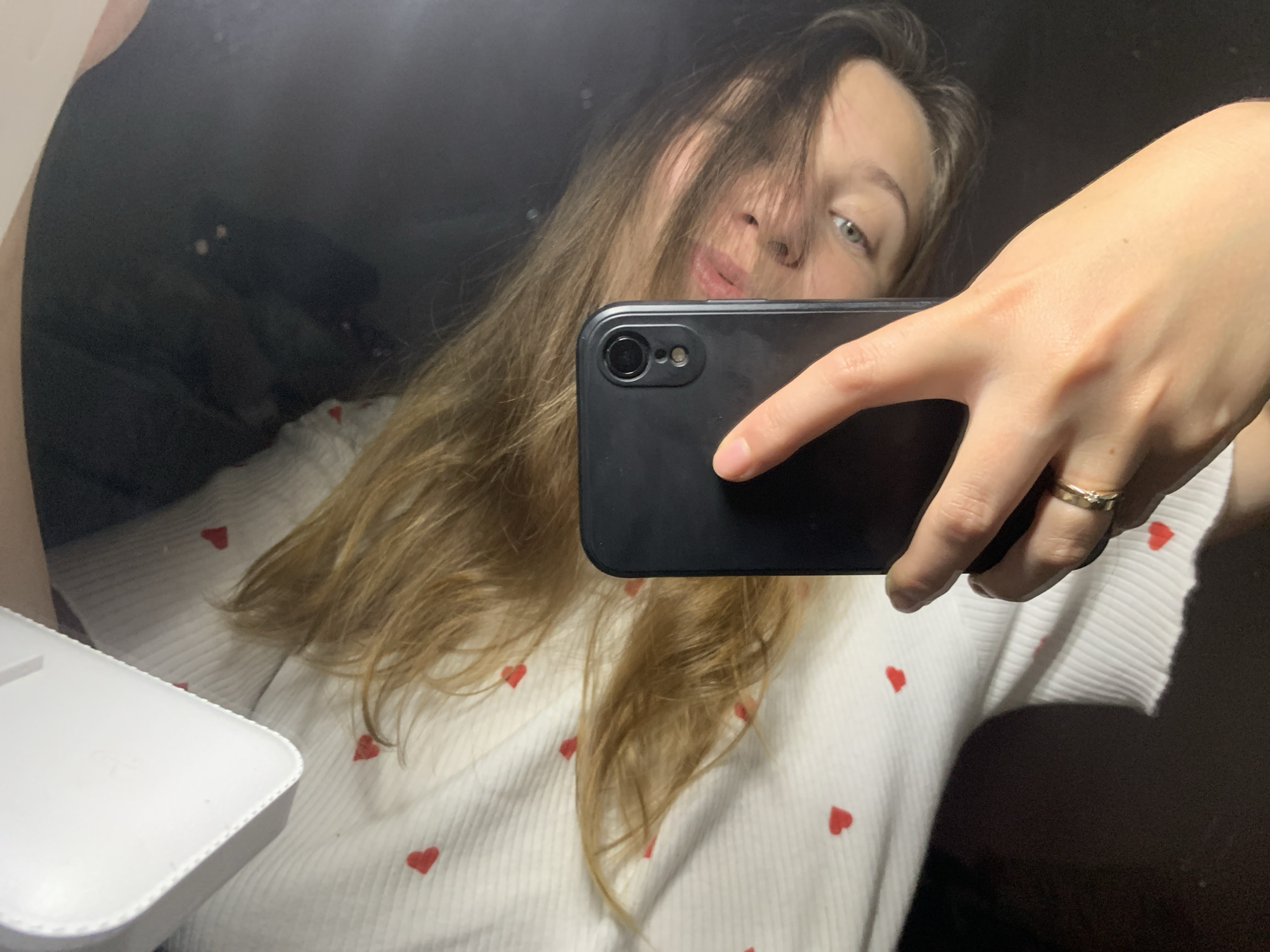 A woman is taking a selfie in a mirror. She is holding a black smartphone with her right hand. She is wearing a white shirt with red hearts. Her hair is light brown and covers part of her face. A white object is visible to the left. The location is Chaikovsky, Russia.